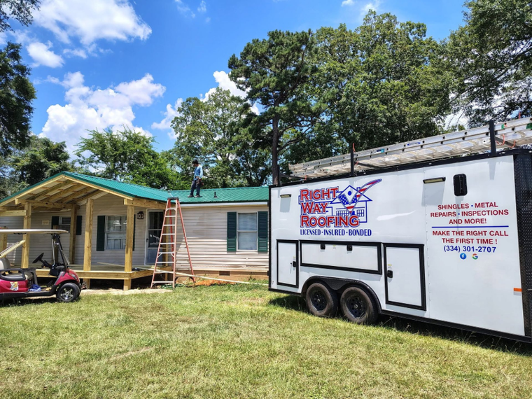 Small house and porch beside a white trailer on a grassy lot under a blue sky