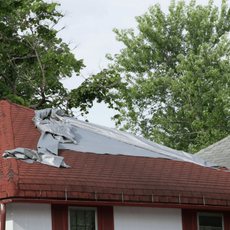Damaged roof with torn gray tarp over red brick house, trees in the background