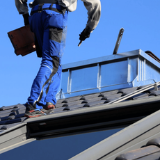 Worker on a rooftop near a skylight, carrying tools and a blue bin under a clear sky