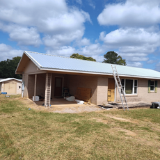 Single-story brick house under construction with a metal roof, ladders, and tools in the yard