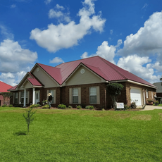 Single-story brick house with a red metal roof and a large green lawn under a partly cloudy blue sky