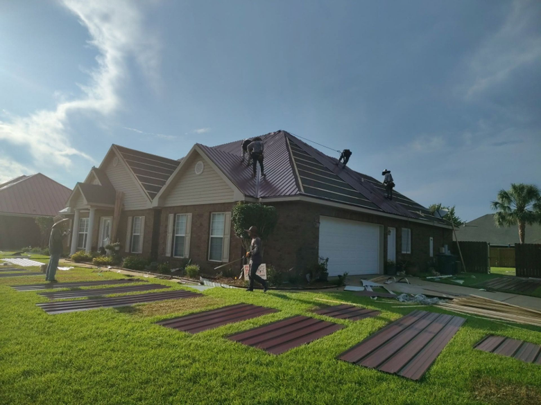 Workers repairing a brick house roof under a sunny sky, with roofing materials on the lawn