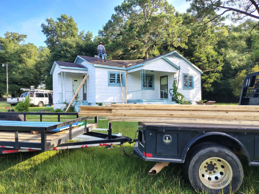 Damaged white house with a person on the roof and a trailer in the foreground amid trees