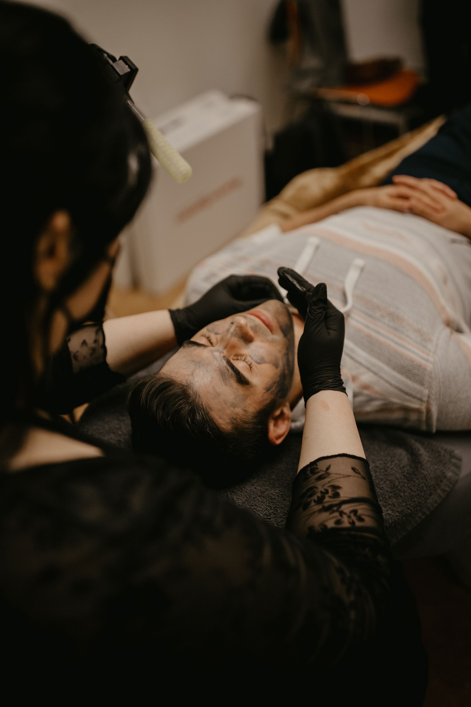 Specialist in black gloves working on a client lying on a couch, in a dim studio