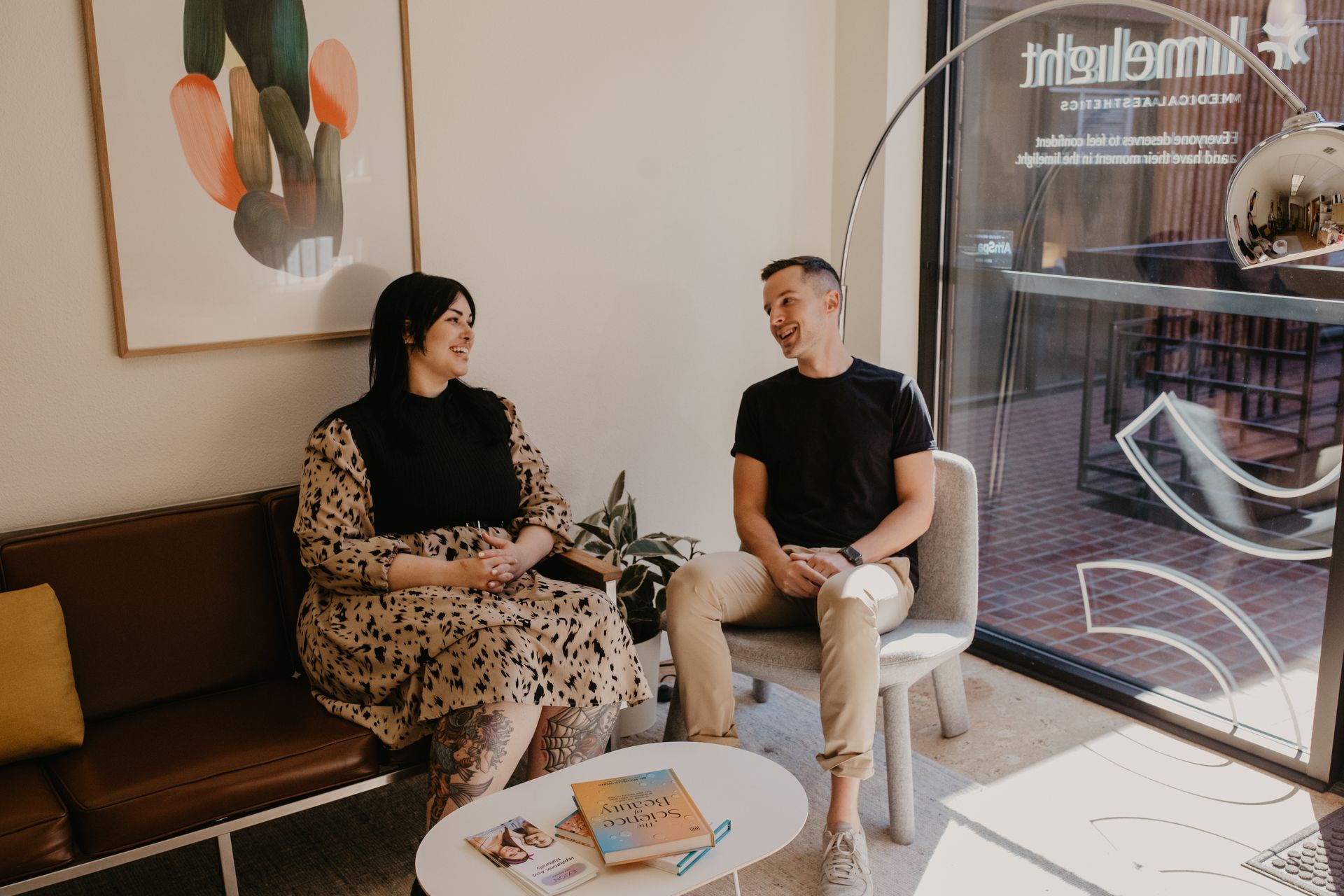Two people chatting in a bright room by a large window, with coffee and a table of brochures.