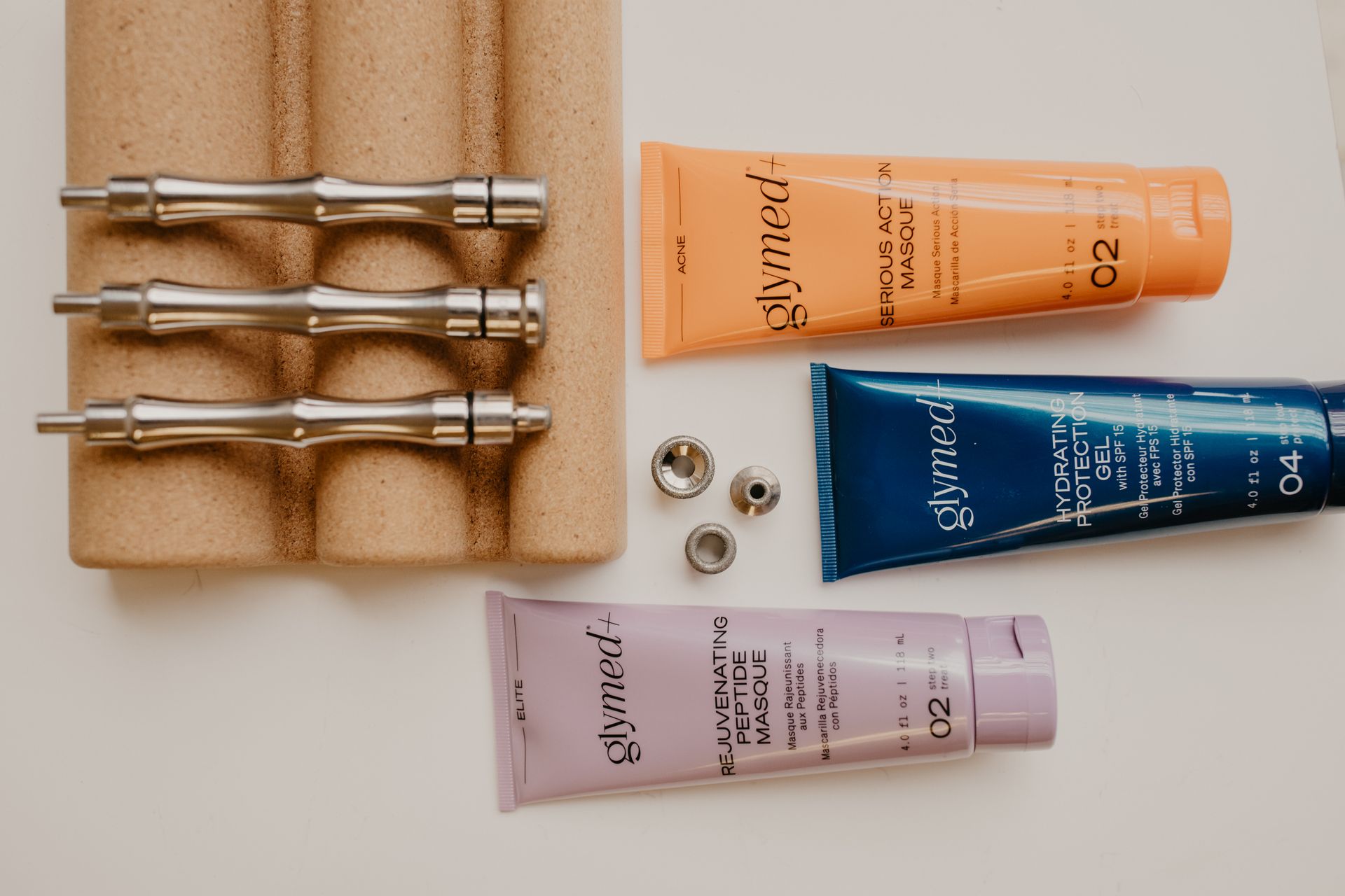 Three silver cosmetic microneedling pens on a cork stand, next to three GlyMed Plus tubes and small metal tips.