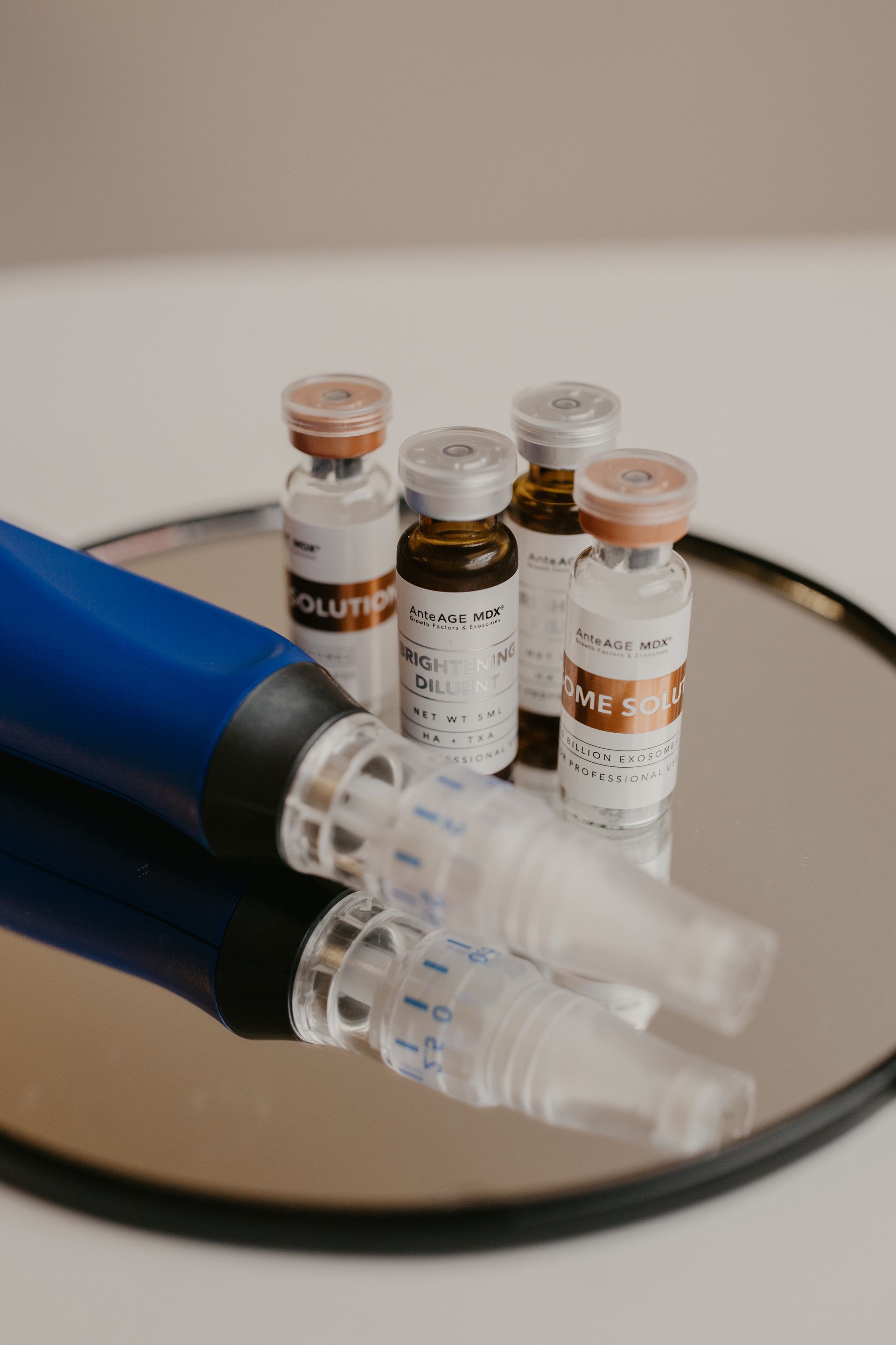 A blue microneedling pen lies on a round mirror next to several small vials used for cosmetic skin treatments.