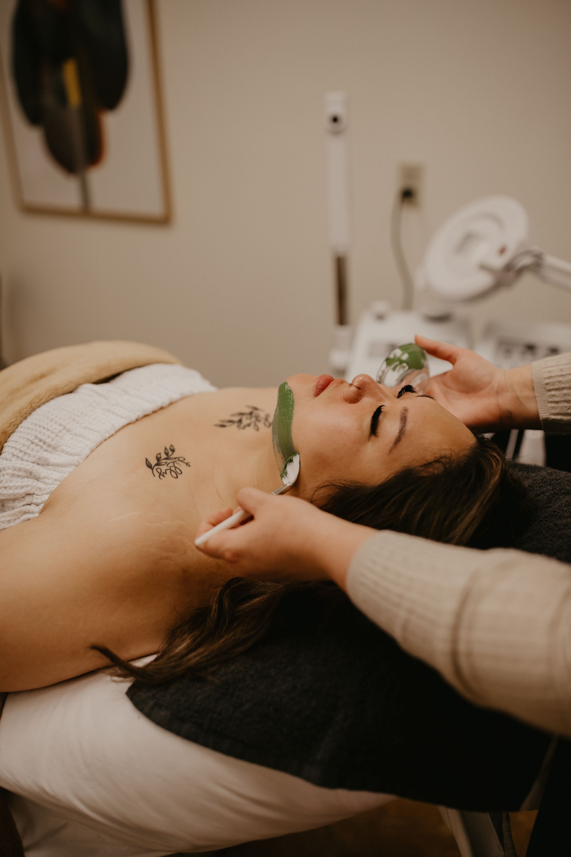 Woman lying on a spa bed while a therapist massages her face, with a towel and warm treatment room.