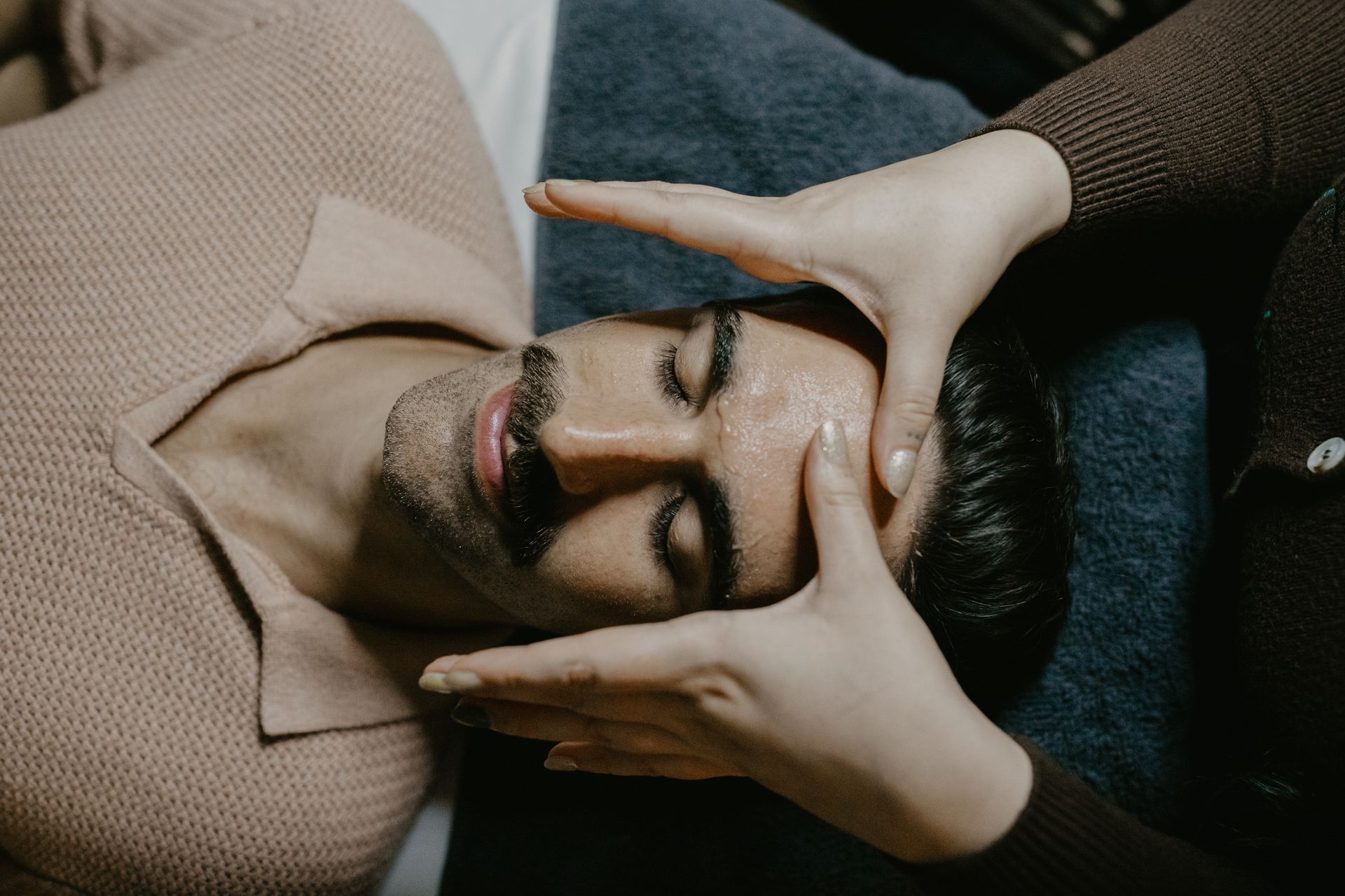 Person lying down with hands framing their face, eyes closed, wearing a beige sweater on a dark couch.