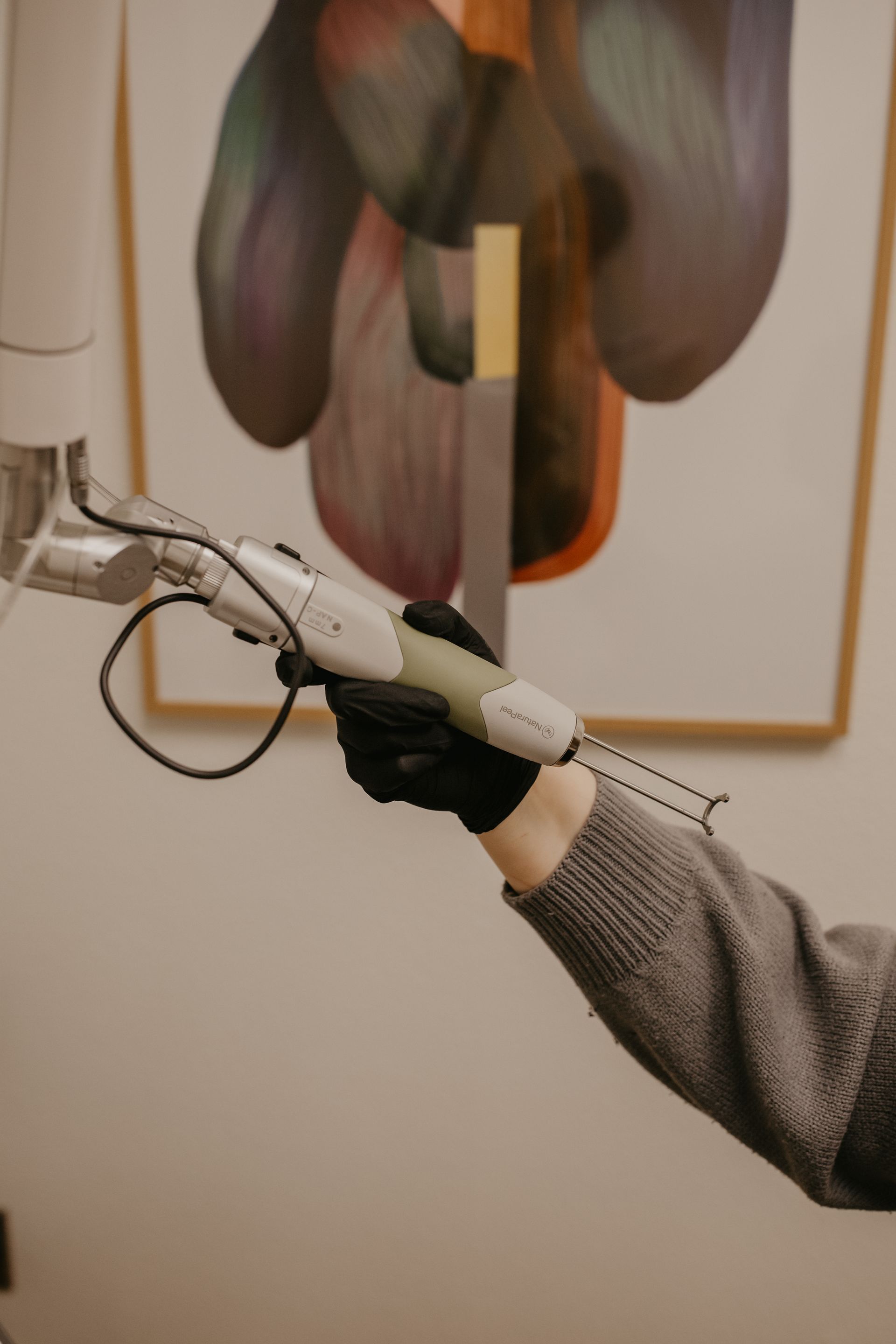 A gloved hand holds a medical laser device, mounted on an articulated arm, in a room with abstract artwork on the wall.