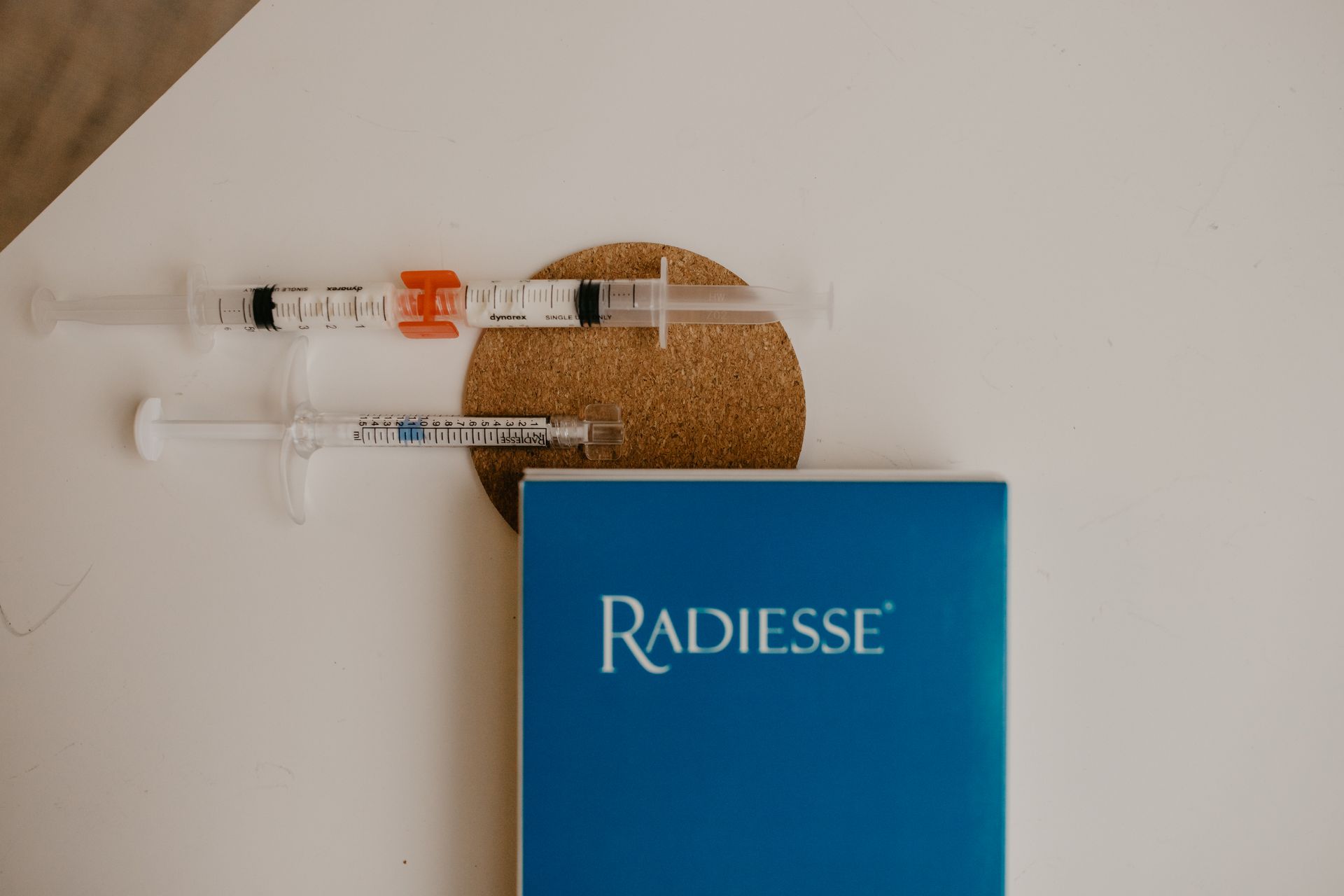 Two medical syringes resting on a cork coaster next to a blue Radiesse product box on a white surface.