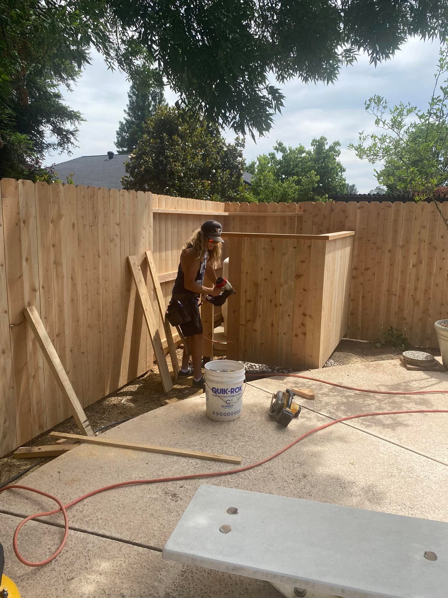 Worker building a wooden fence and shed in a backyard, with tools and lumber on the ground.