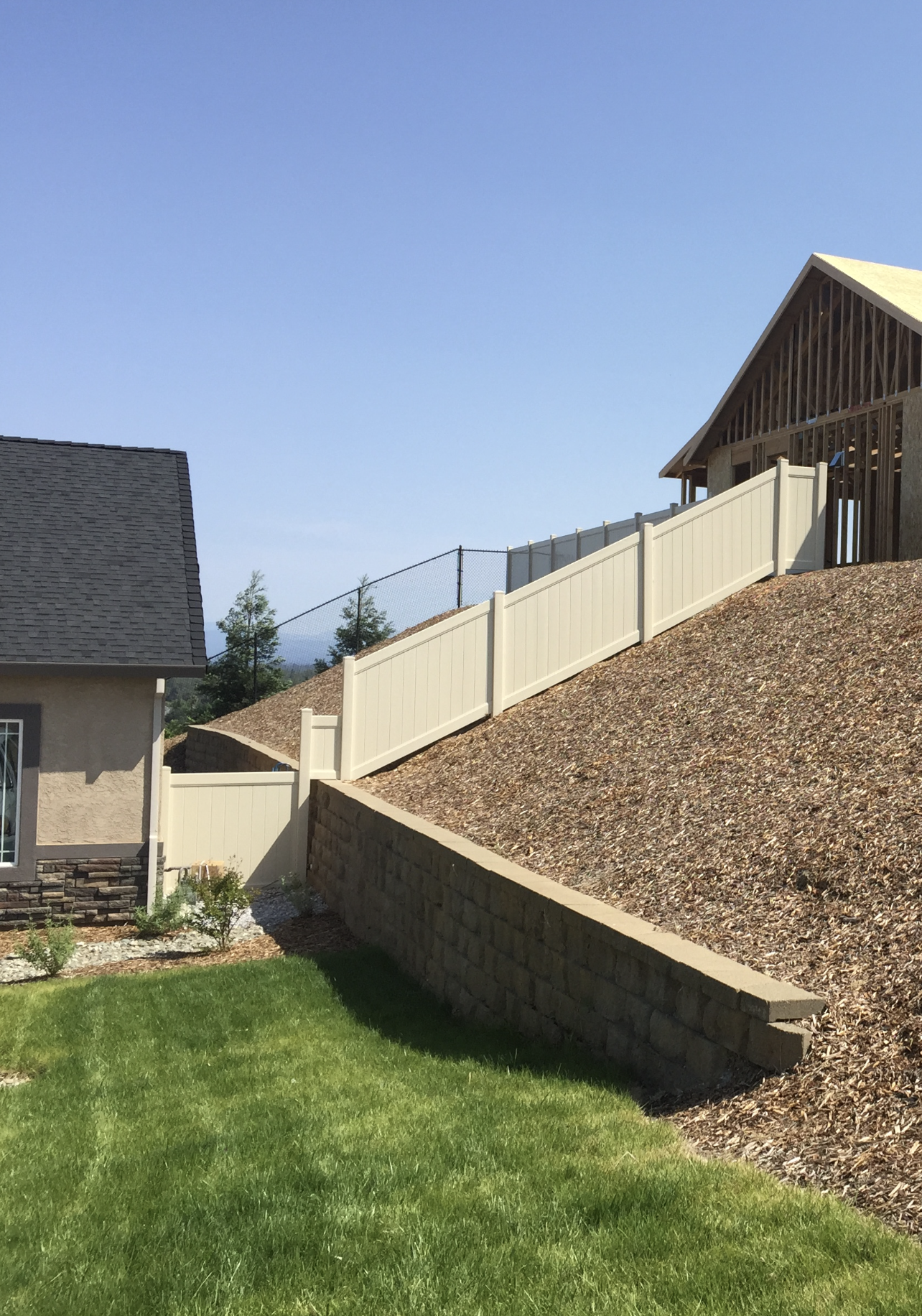Backyard with green lawn, sloped stone retaining wall, and beige fence under a clear blue sky