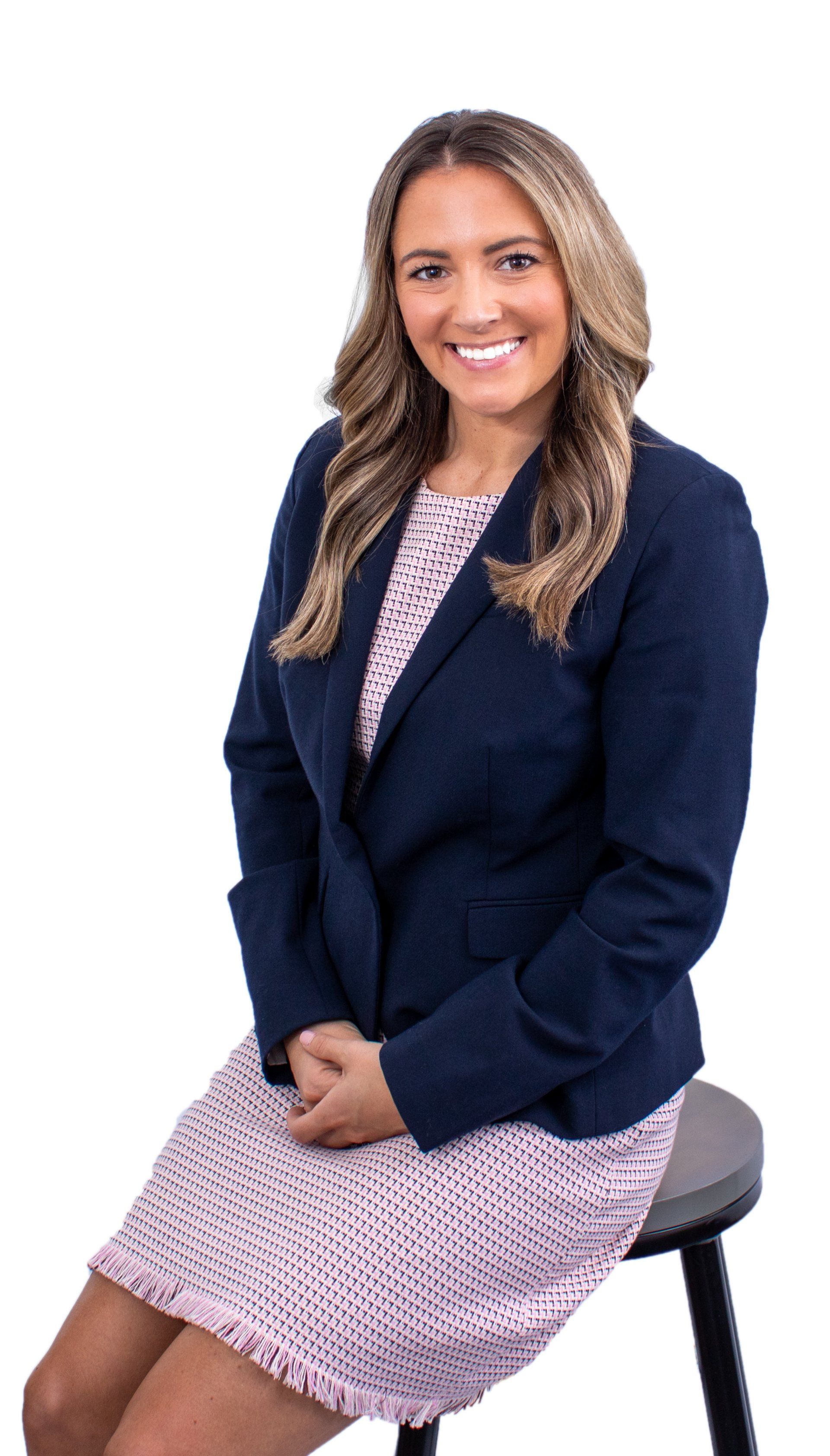 Woman in blazer and patterned dress, smiling, seated on a stool.
