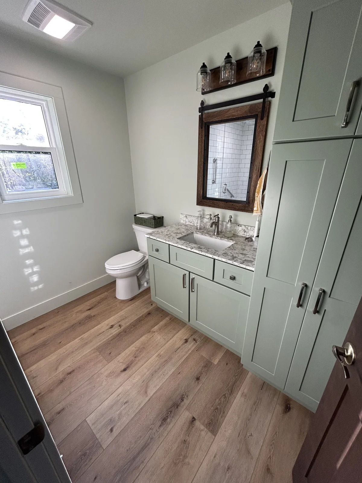A bathroom with sage green cabinets, wood-look flooring, a sink, a toilet, and a window with a barn-style wood mirror.