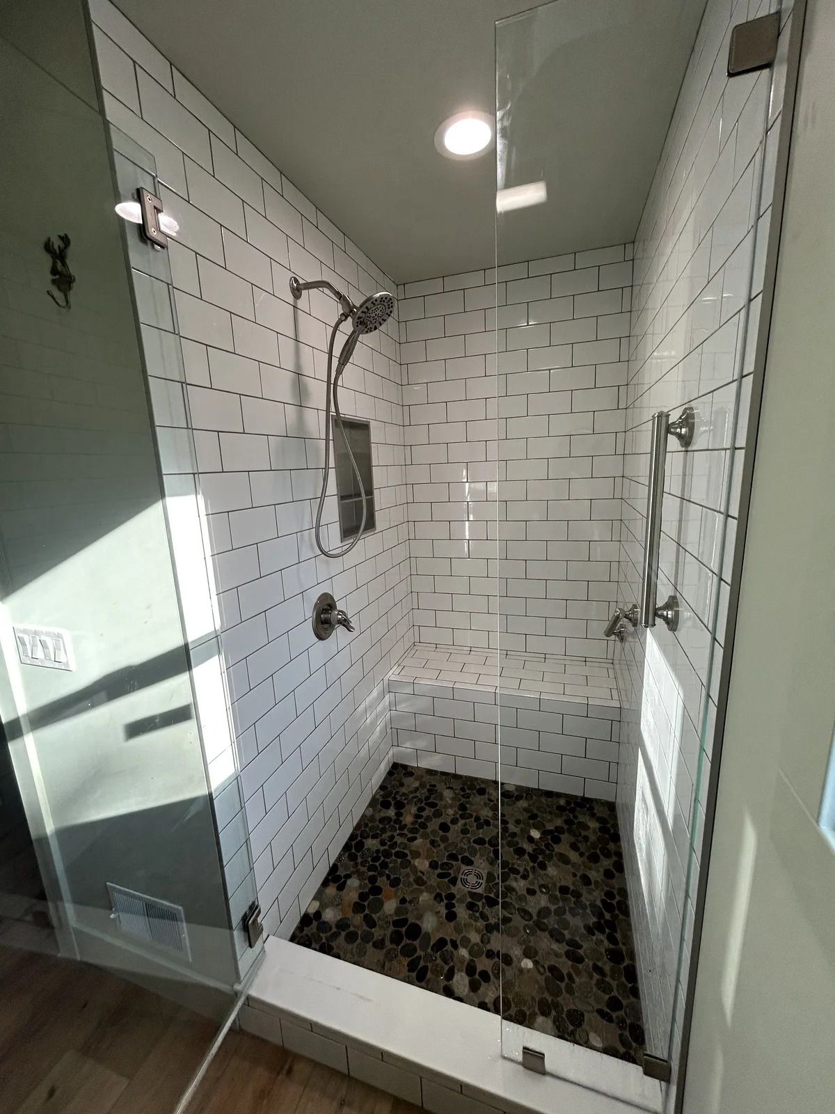 A modern walk-in shower with white subway tile walls, a pebble floor, a built-in bench, and a glass door.