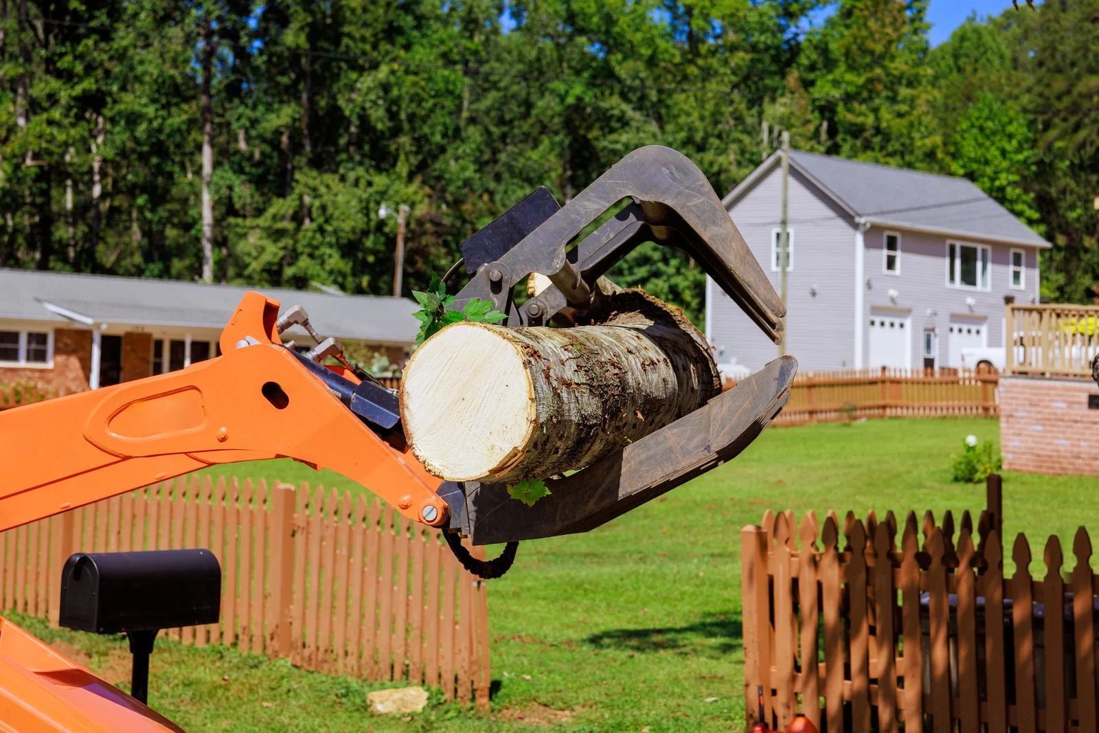 Excavator gripping a large log in a suburban yard beside a wooden fence and house