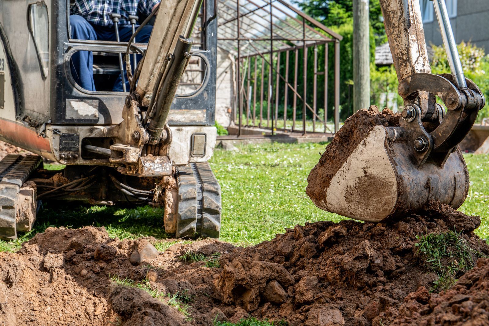 Mini-excavator digging a trench in a grassy yard, near a fence and a person in the cabin.
