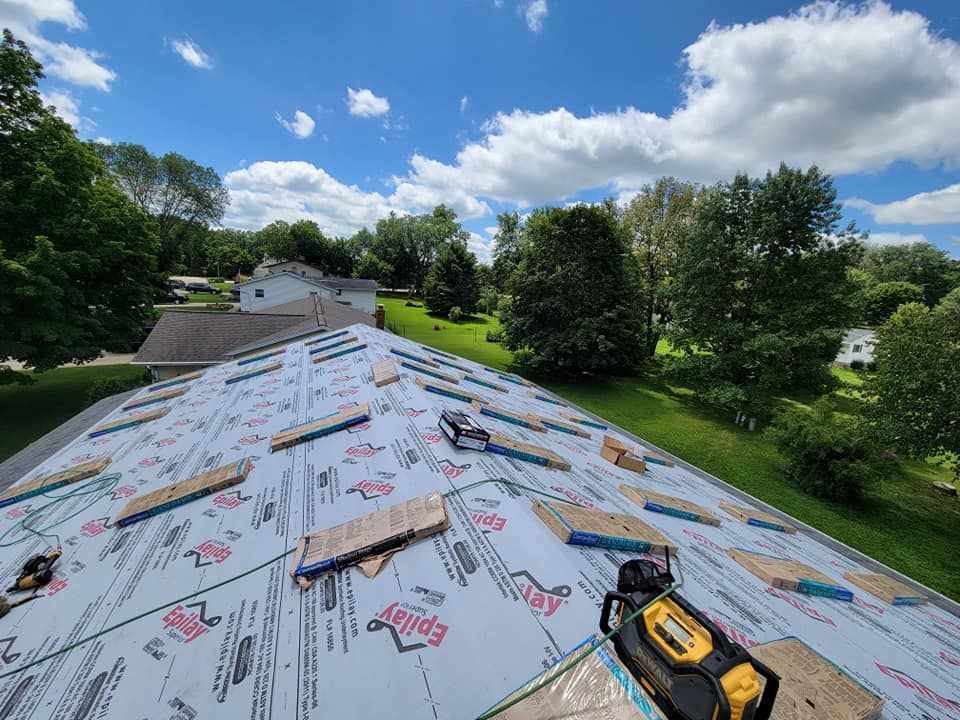 Roof under construction with gray underlayment, scattered tools, and trees under a blue sky