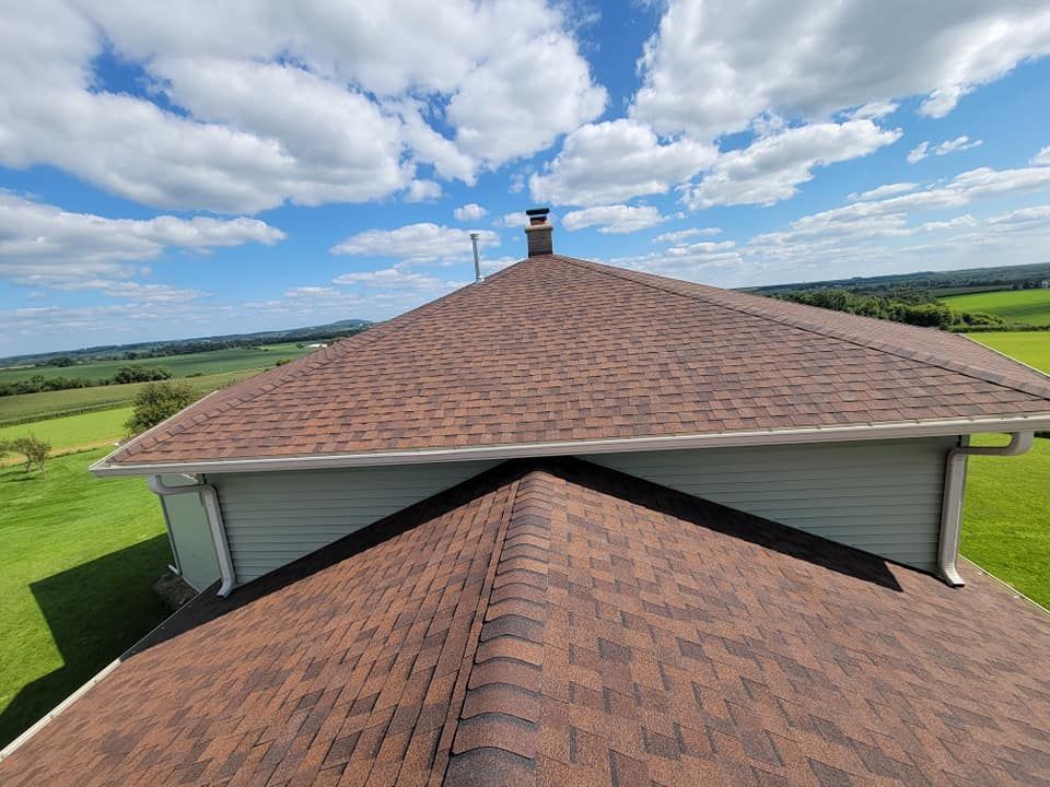 View over a brown shingled roof with a small cupola, overlooking green fields and a cloudy blue sky.
