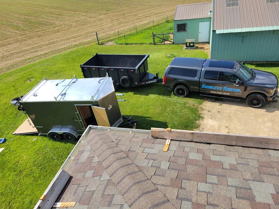 Blue pickup, black trailer, and metal shed beside a farm building and tilled field.