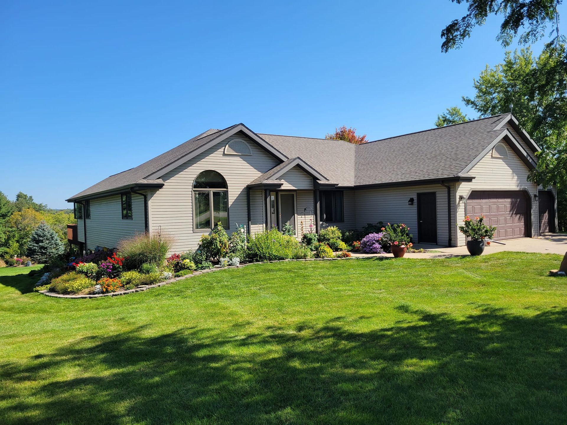 Single-story suburban house with gray siding, landscaped flower beds, and a green lawn under a blue sky