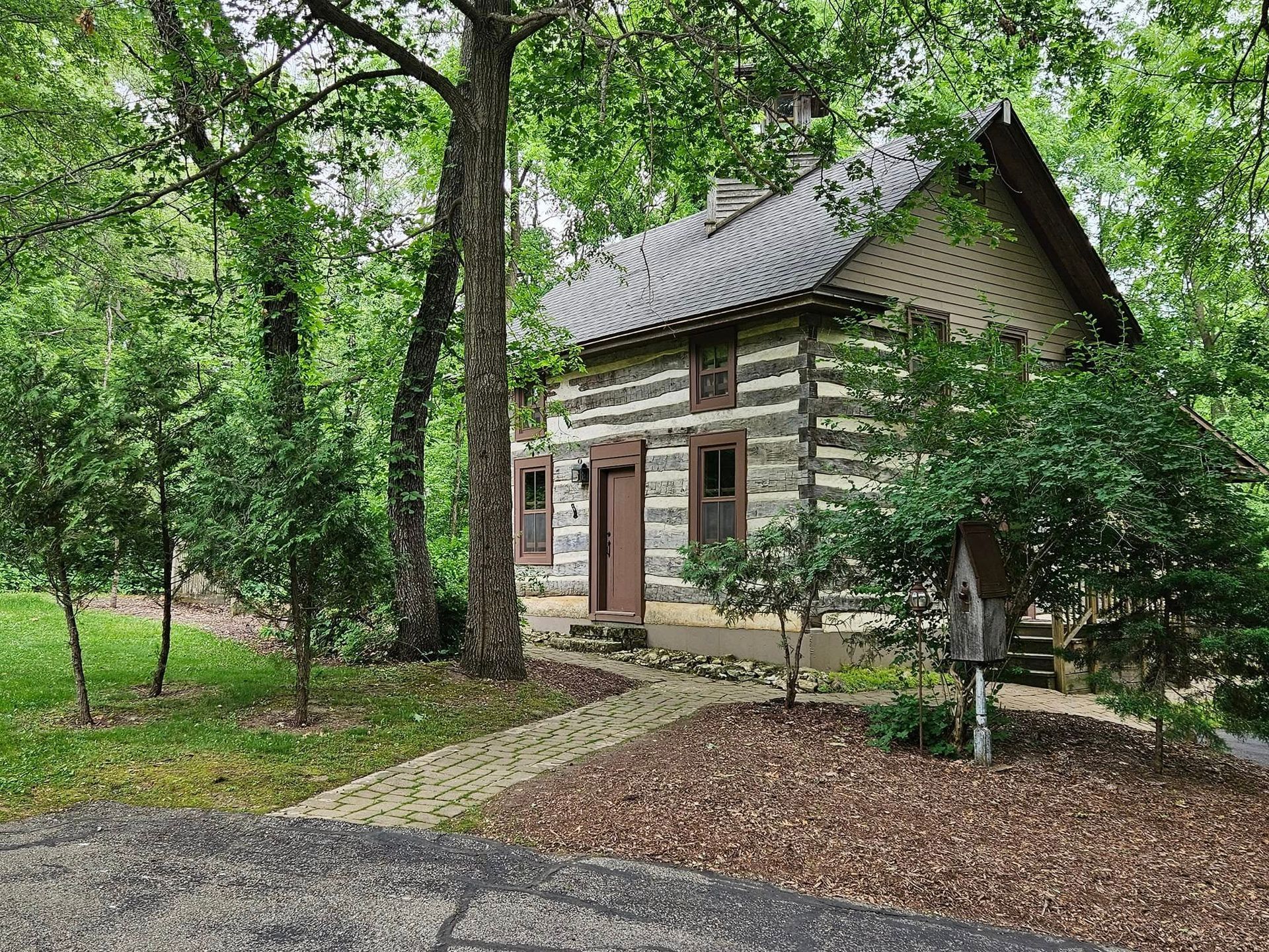 Small log cabin in a leafy forest clearing with a gravel path and tall trees.