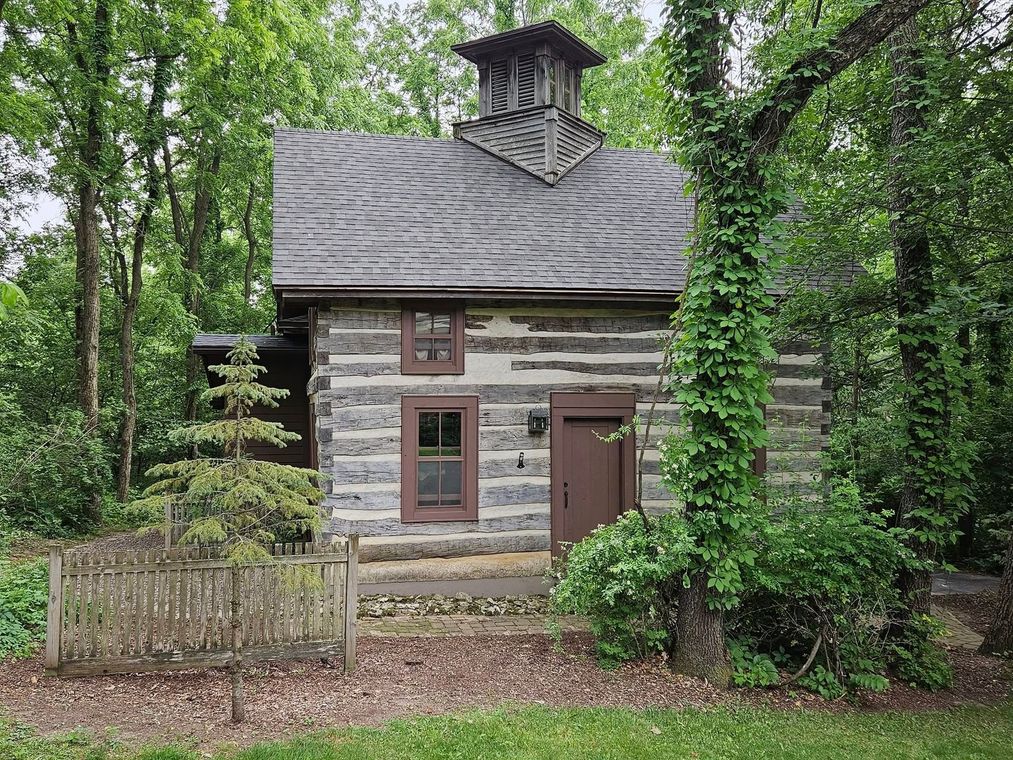 Small rustic log cabin with a wavy roof and chimney, set among green trees and shrubs.