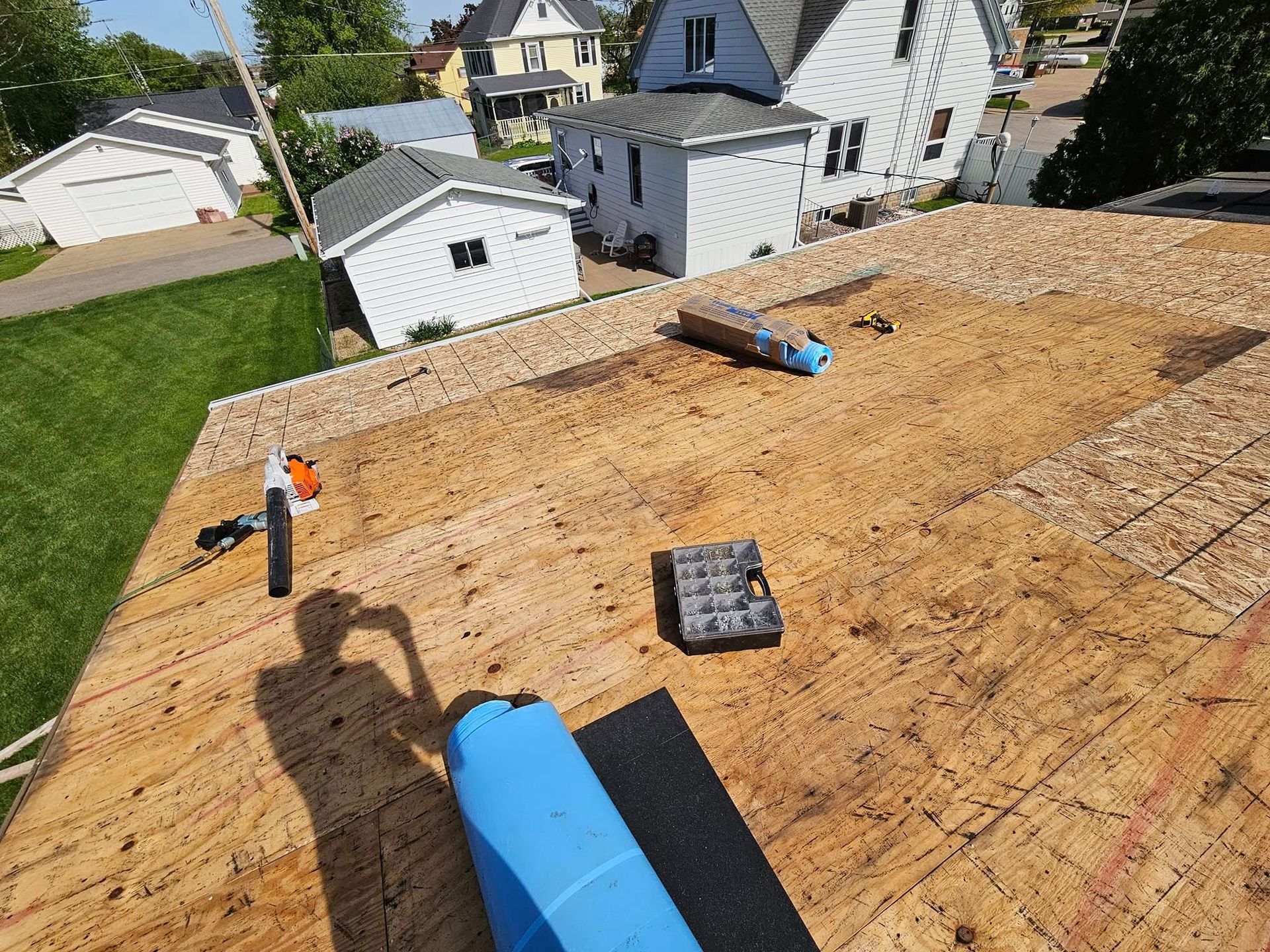 Rooftop under construction with plywood sheathing, blue vent pipes, and nearby houses in view