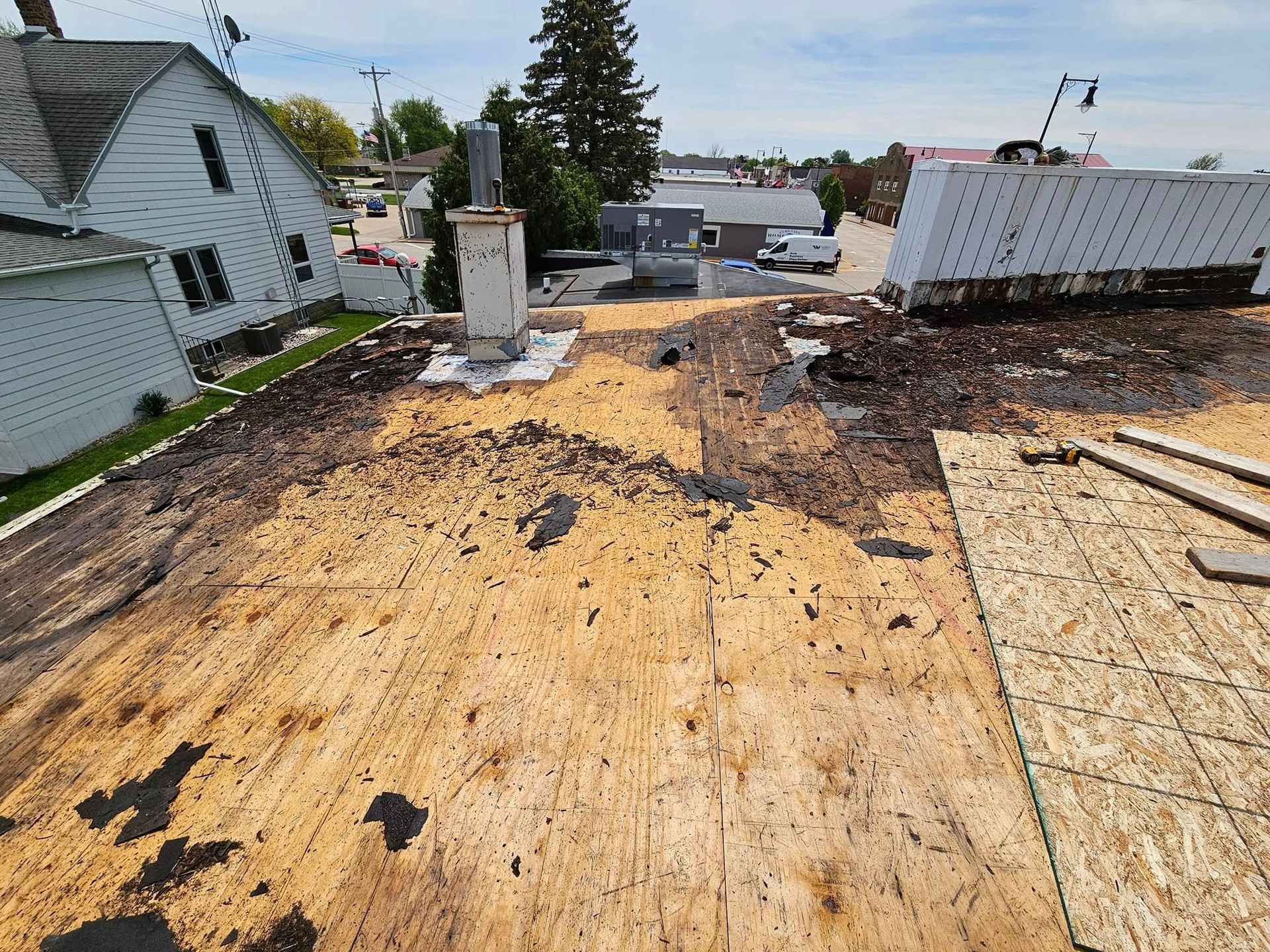 Rooftop under construction with plywood sheathing, debris, and neighboring houses visible on both sides.