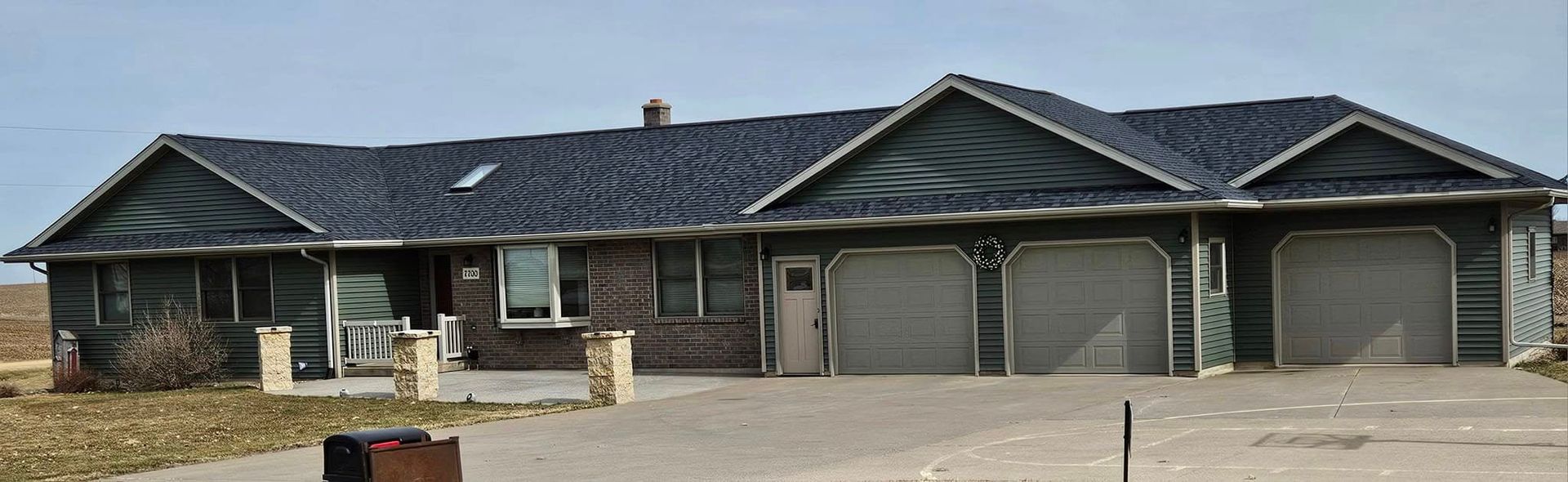 Single-story suburban house with dark roof, stone facade, and three garage doors under a cloudy sky