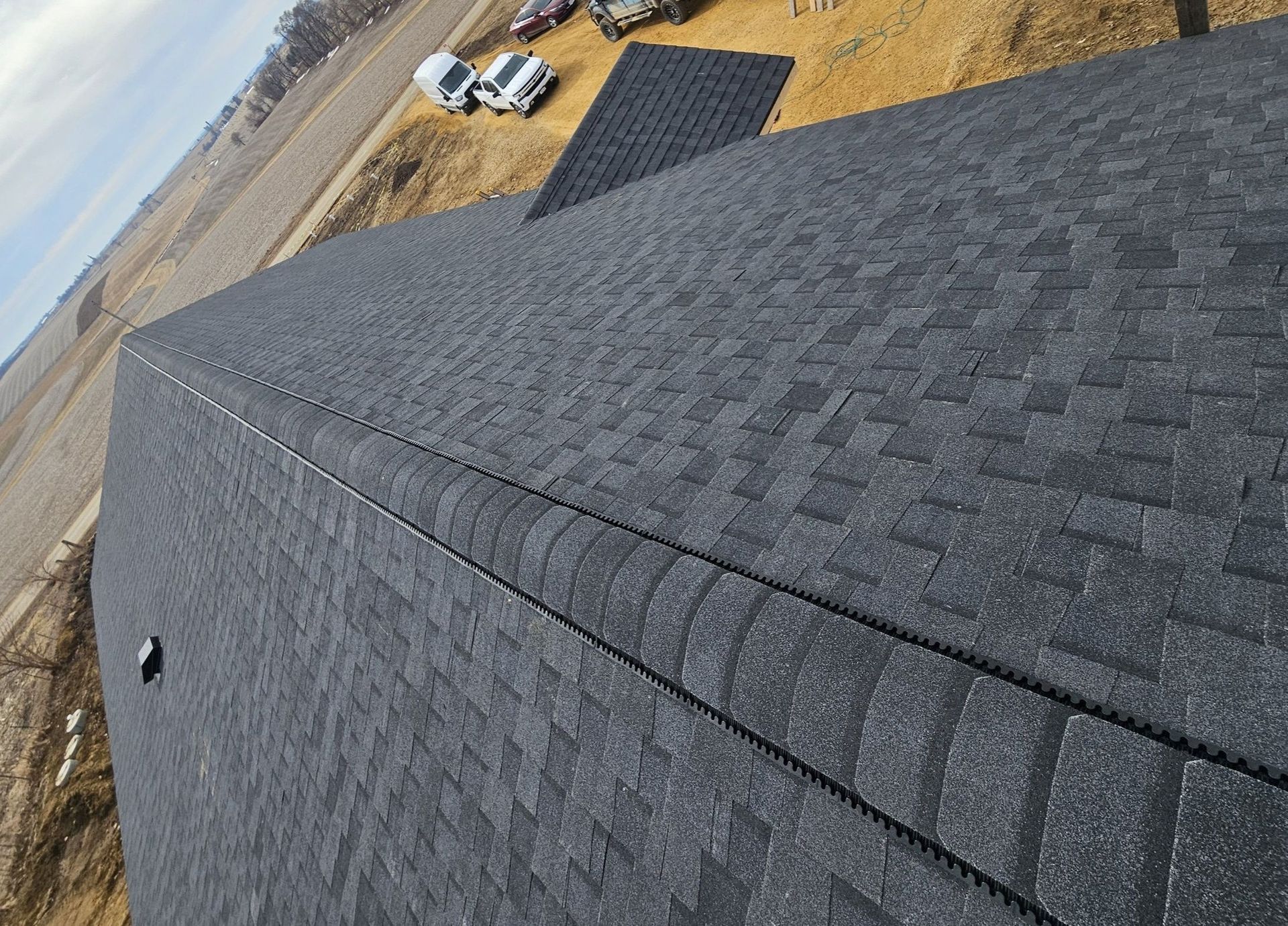 Close-up of a dark gray shingled roof with a road and two white vehicles in the background.