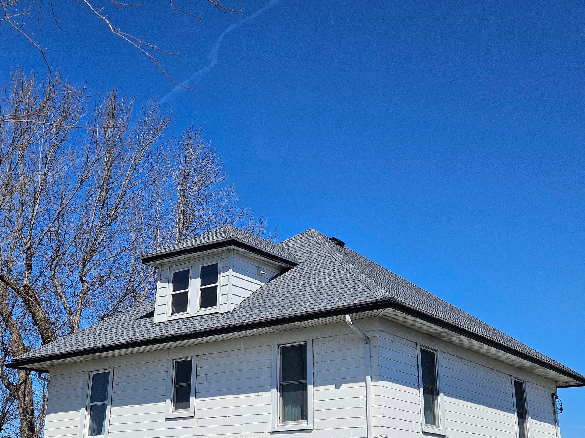 White house with gray roof under a clear blue sky, viewed from below with bare trees nearby