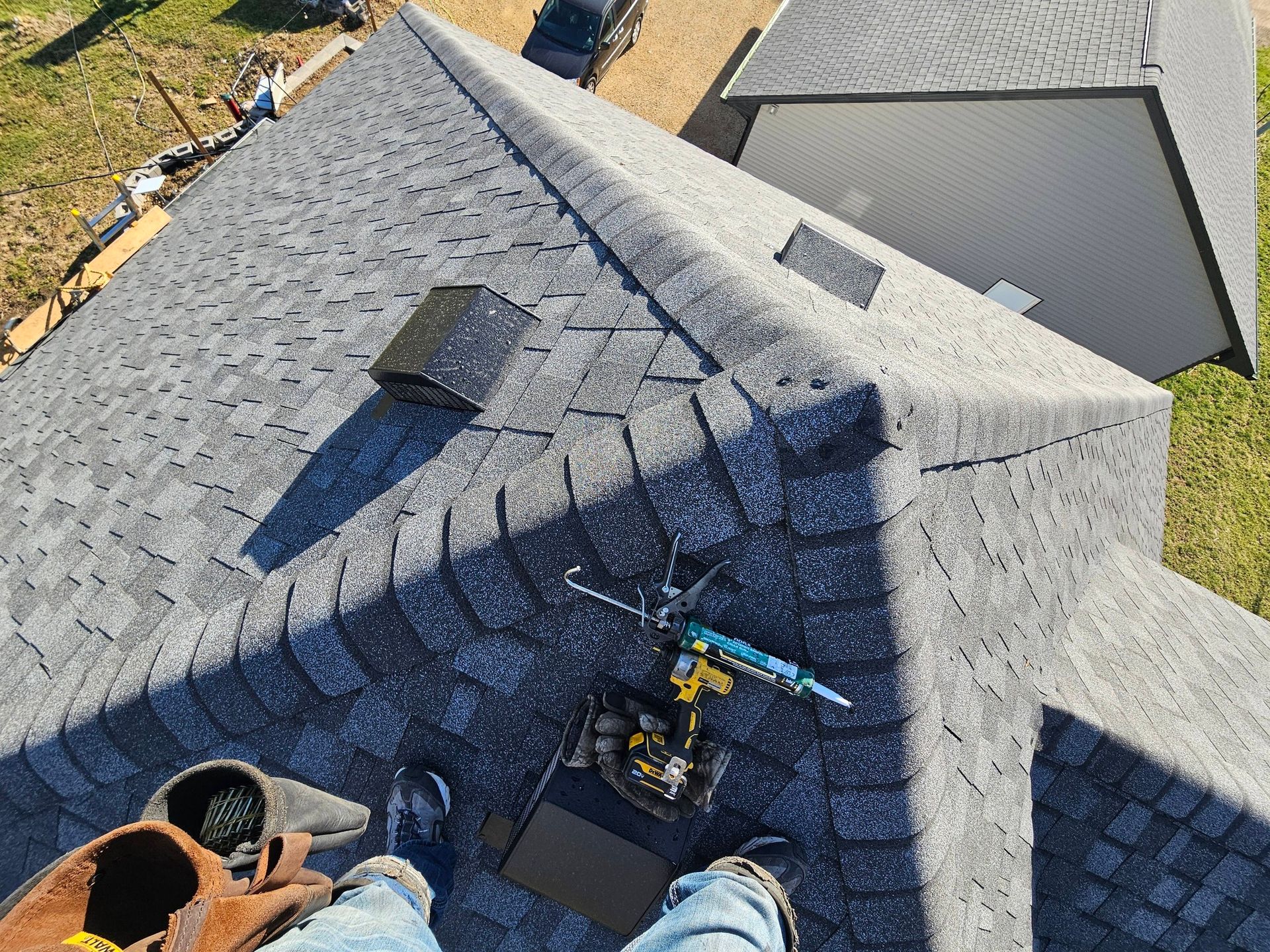 Roofing worker on a gray shingled roof, using tools near a steep ridge and skylight.