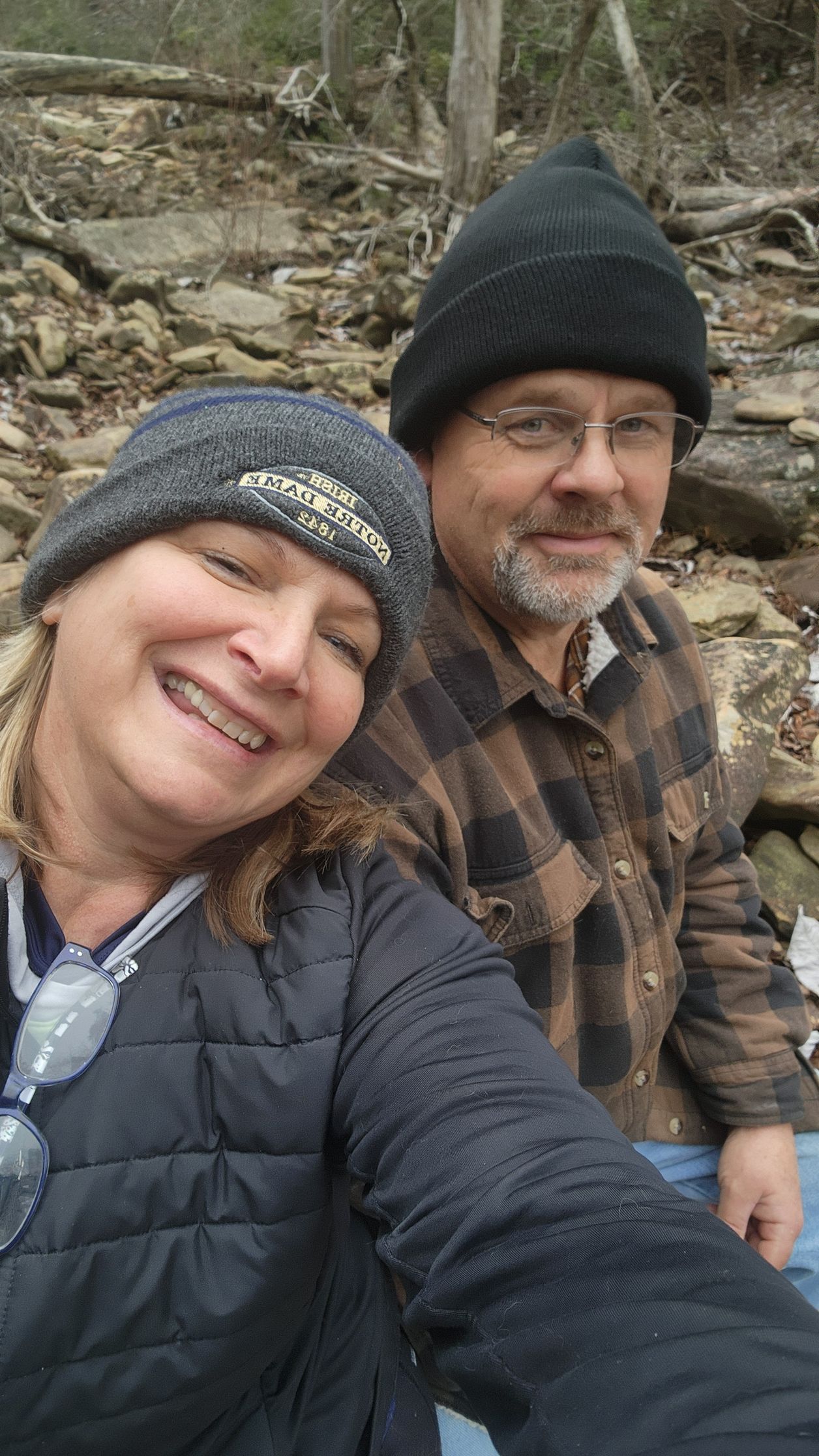 Two smiling people in winter hats and jackets posing outdoors by a rocky stream.