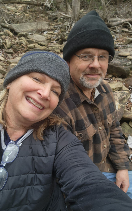 Smiling couple in beanies and winter jackets taking a selfie on a rocky trail.