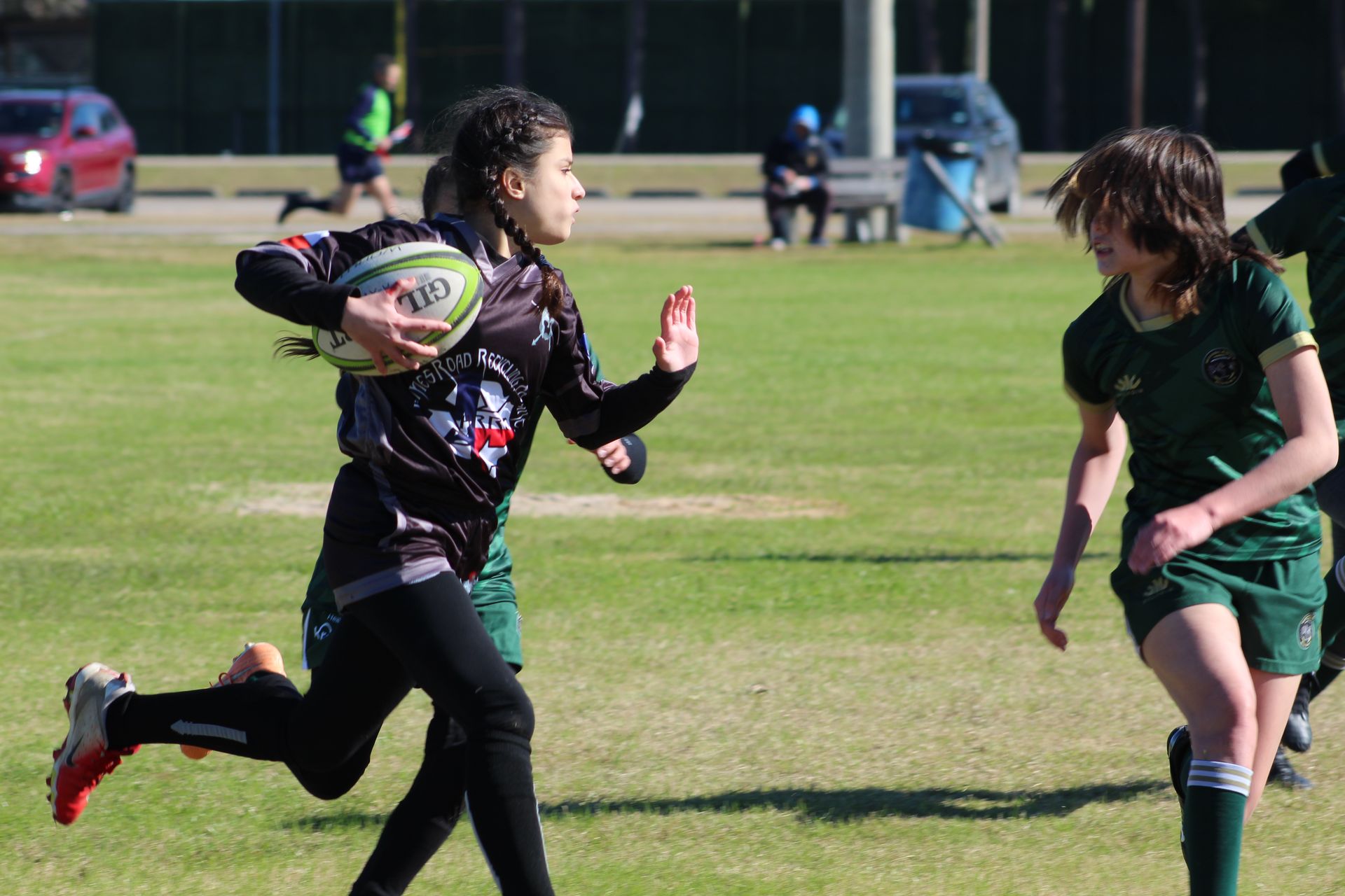 Player in black jersey runs with rugby ball, pursued by player in green jersey on grassy field.