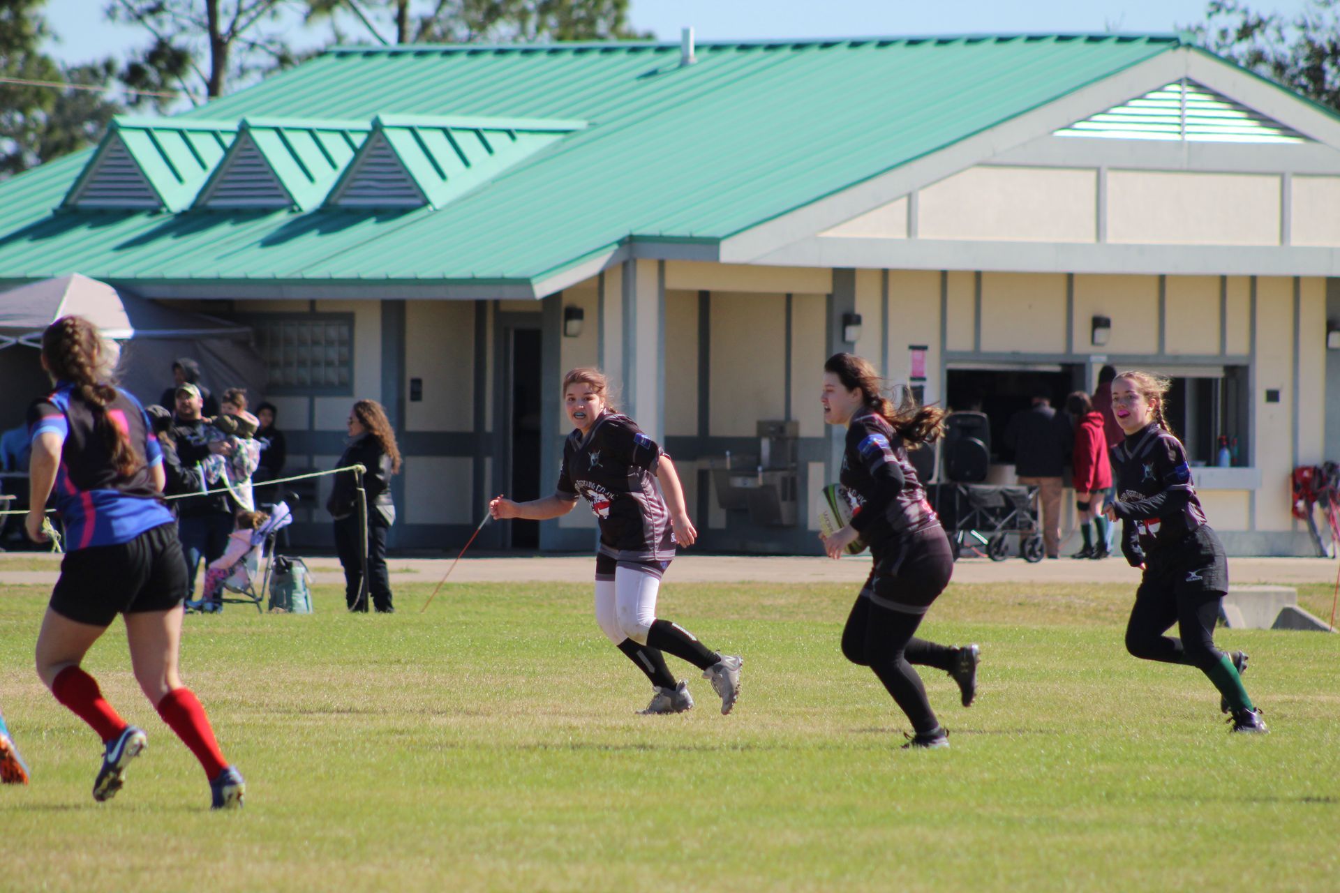Women's rugby game in progress on a grassy field; players in black and red uniforms running toward a building.