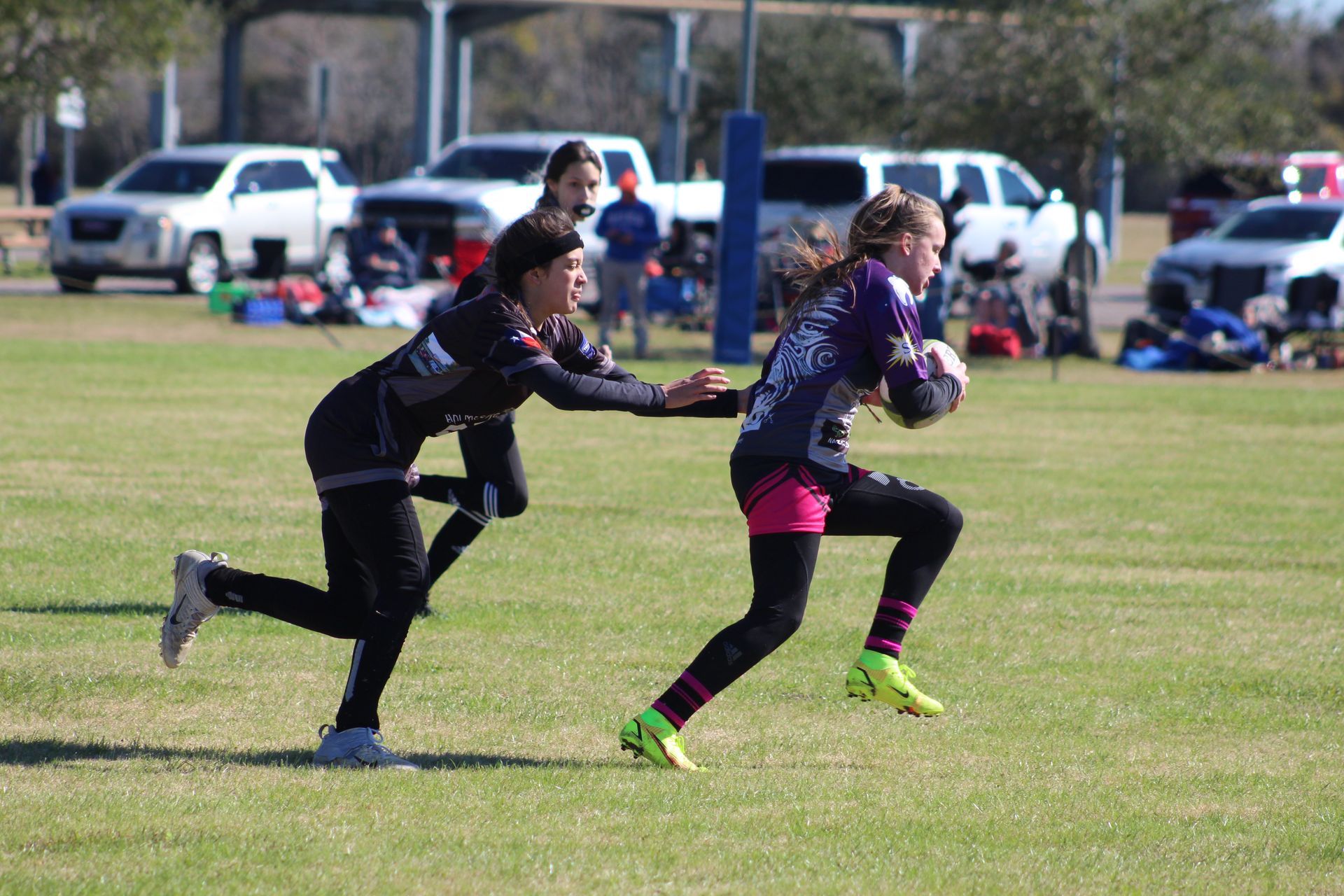 Girl in purple jersey runs with ball; another player attempts to tackle on a grassy field.