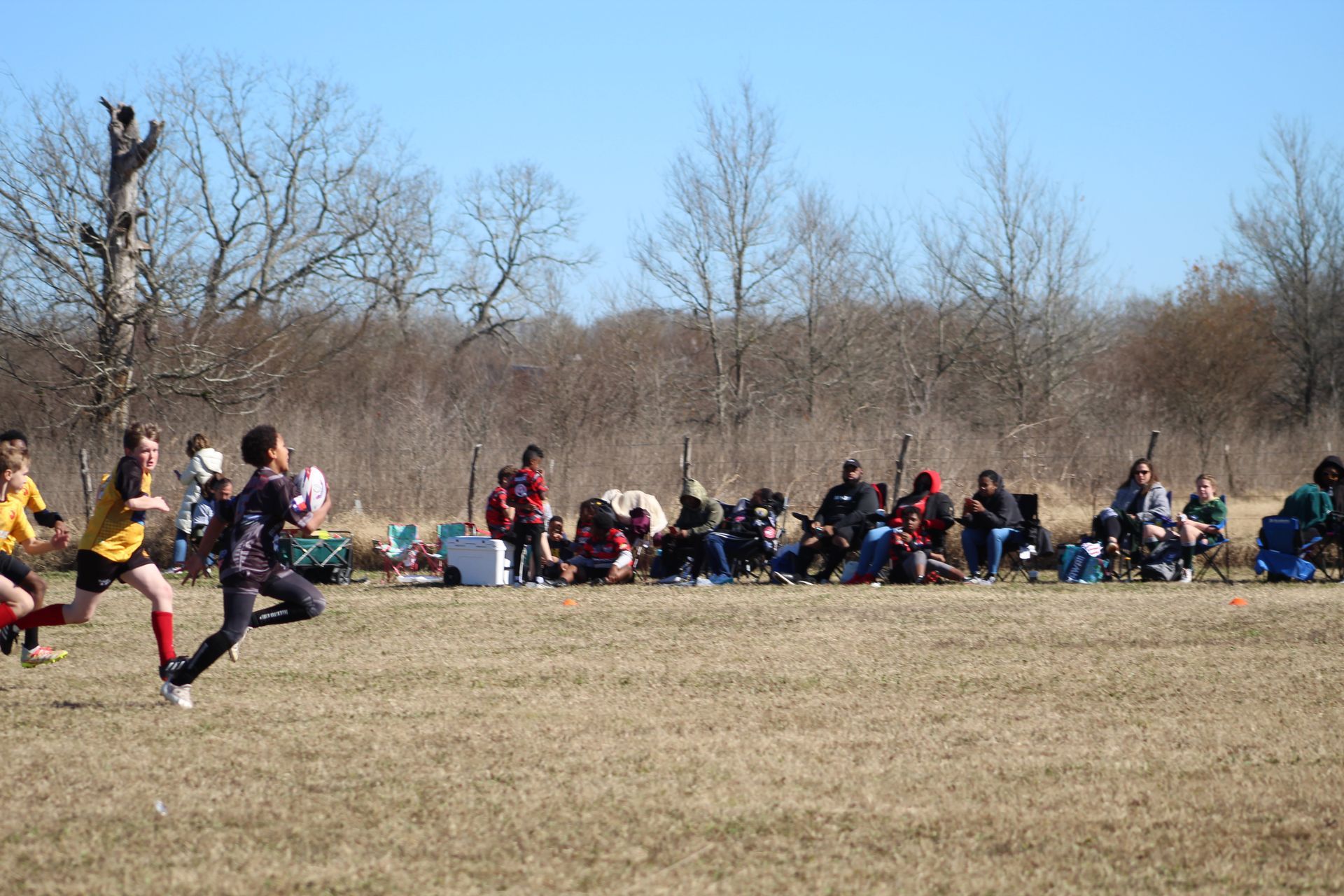 Soccer game in a field: players running, spectators watching from chairs, trees in the background under a blue sky.