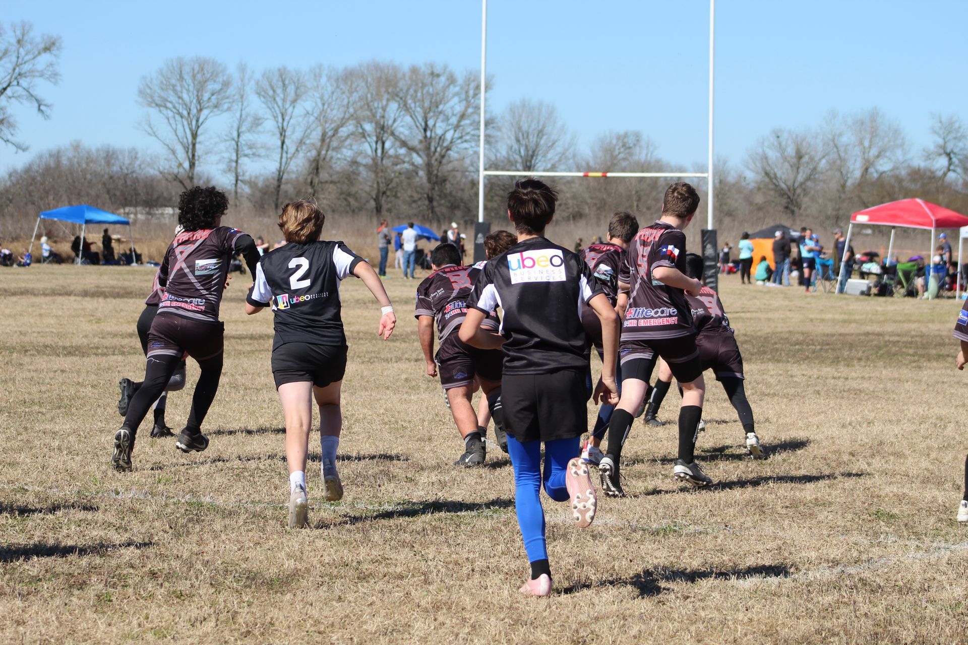 Rugby players in black and pink jerseys running toward goal posts on a sunny field.