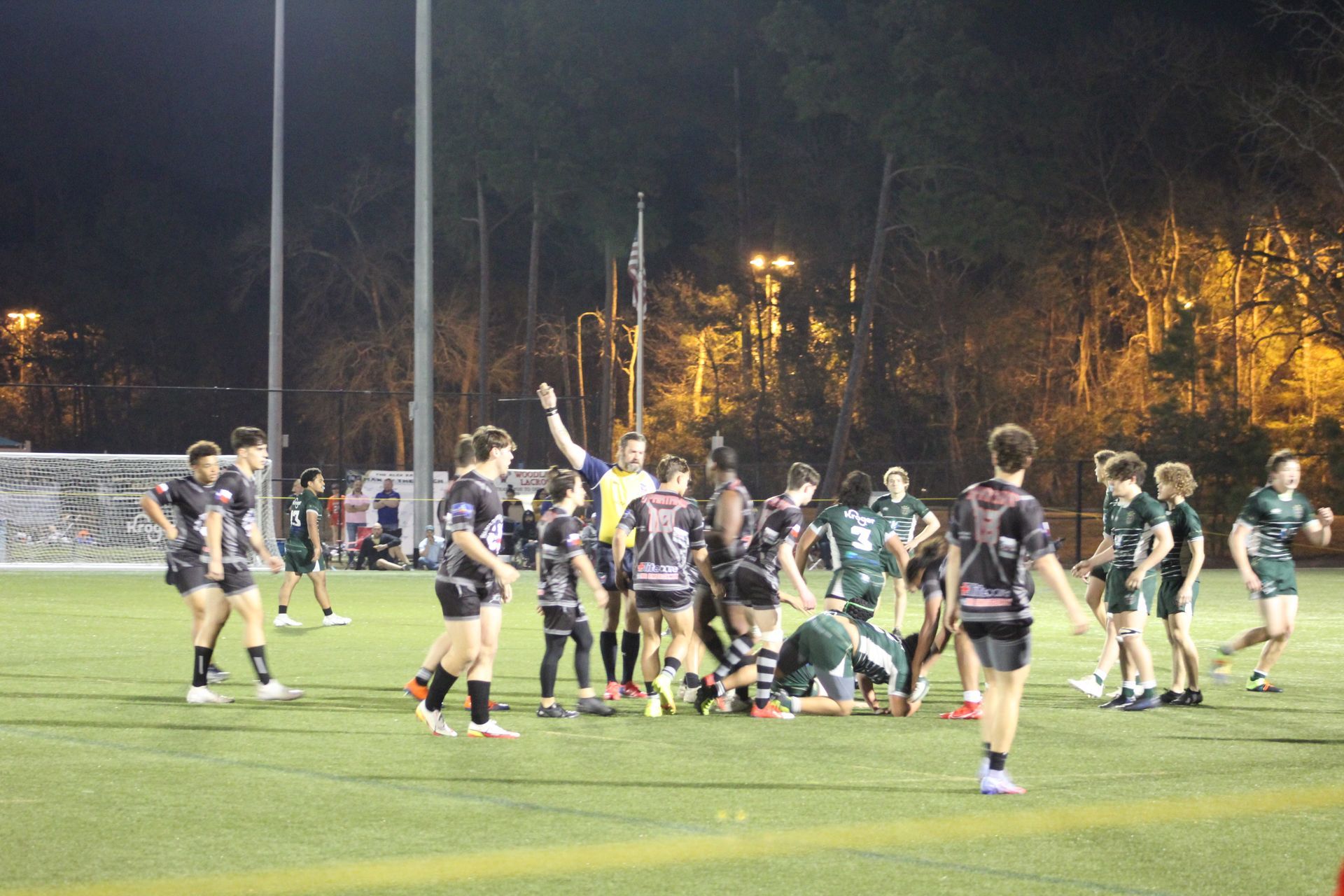 Rugby players on a green field at night, a referee raises their arm; some players in black and white uniforms,