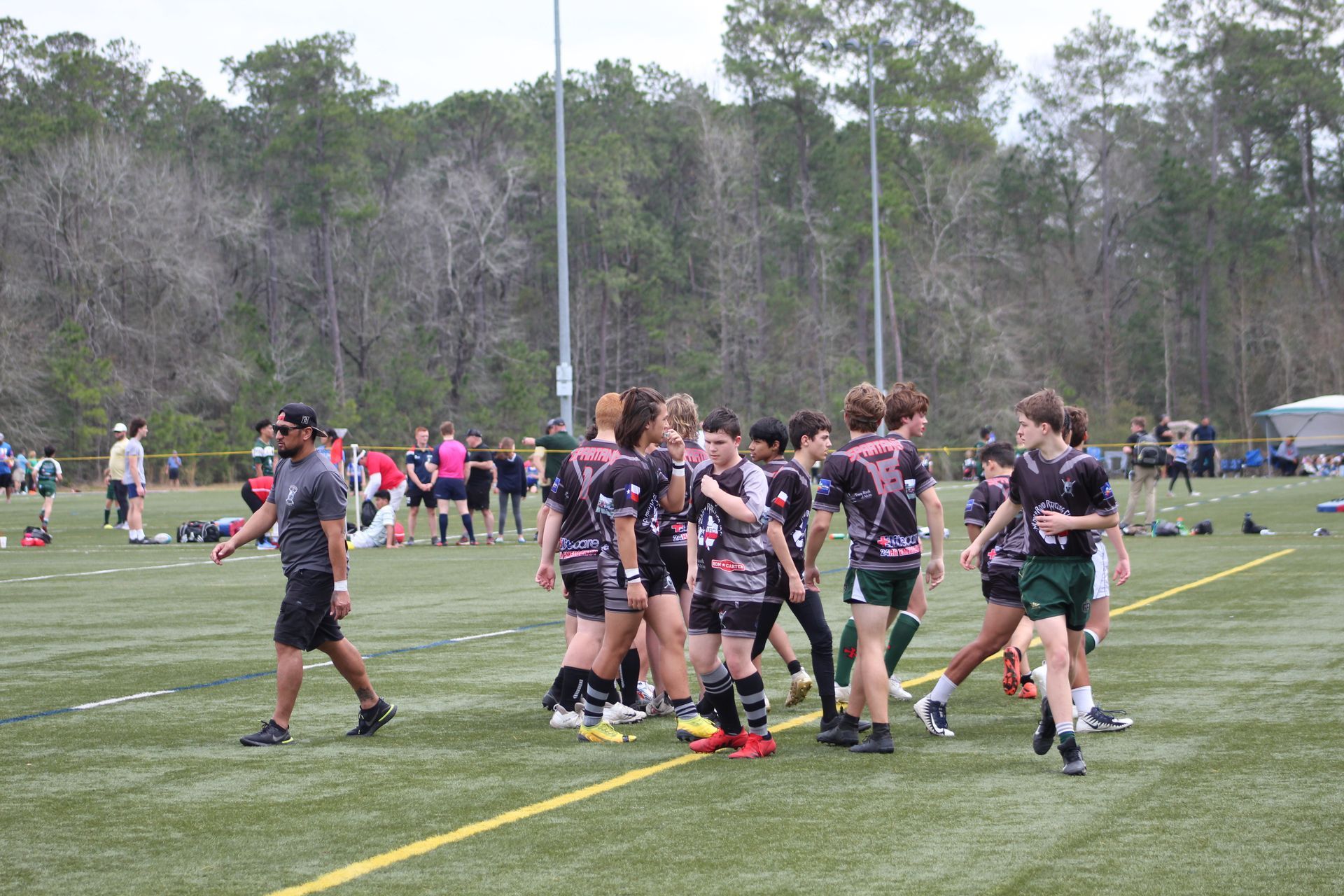 Rugby players in black and pink jerseys huddle on a green field. A coach walks nearby.