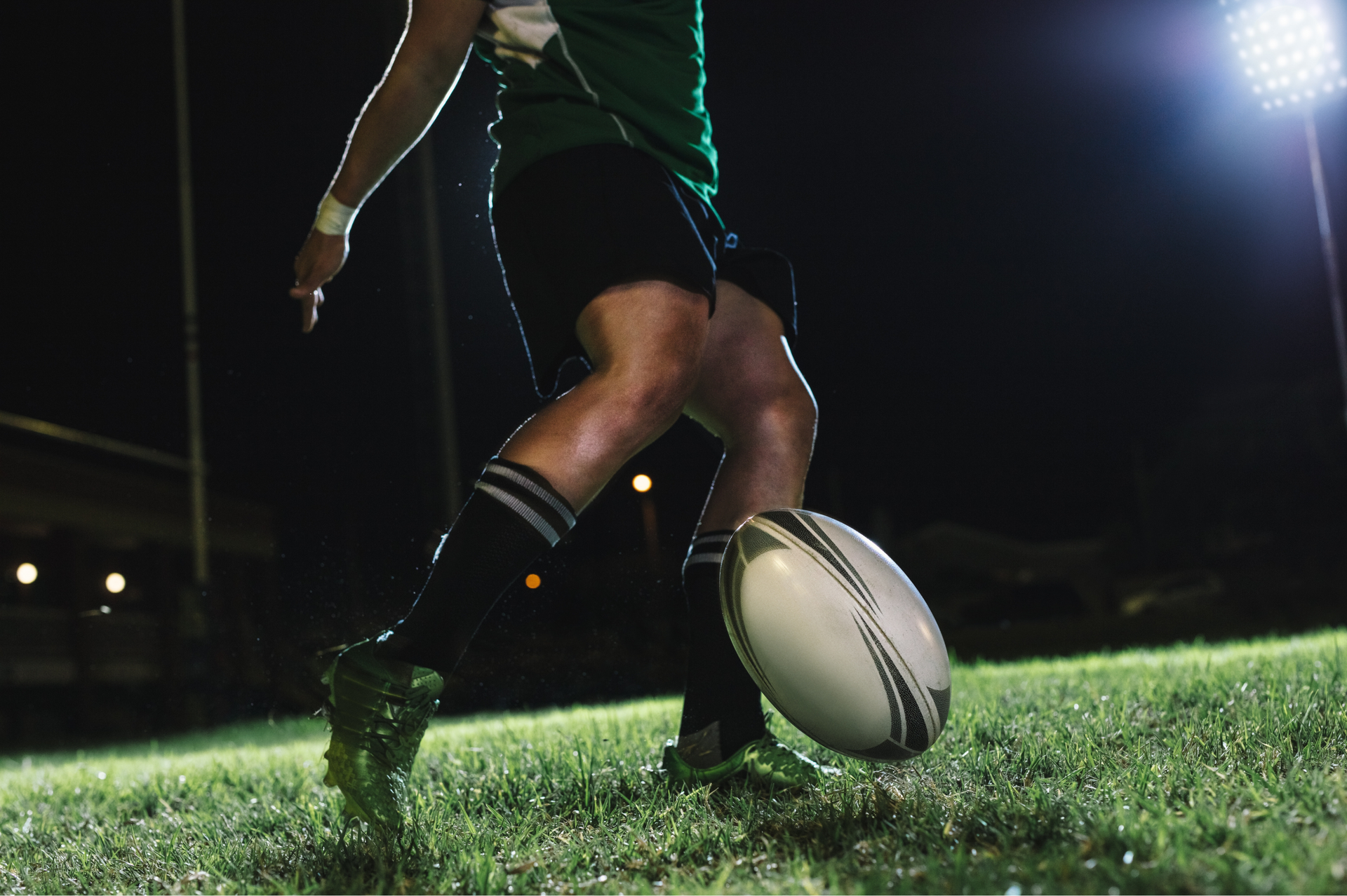 Rugby player kicking oval ball on a green field at night, illuminated by stadium lights.