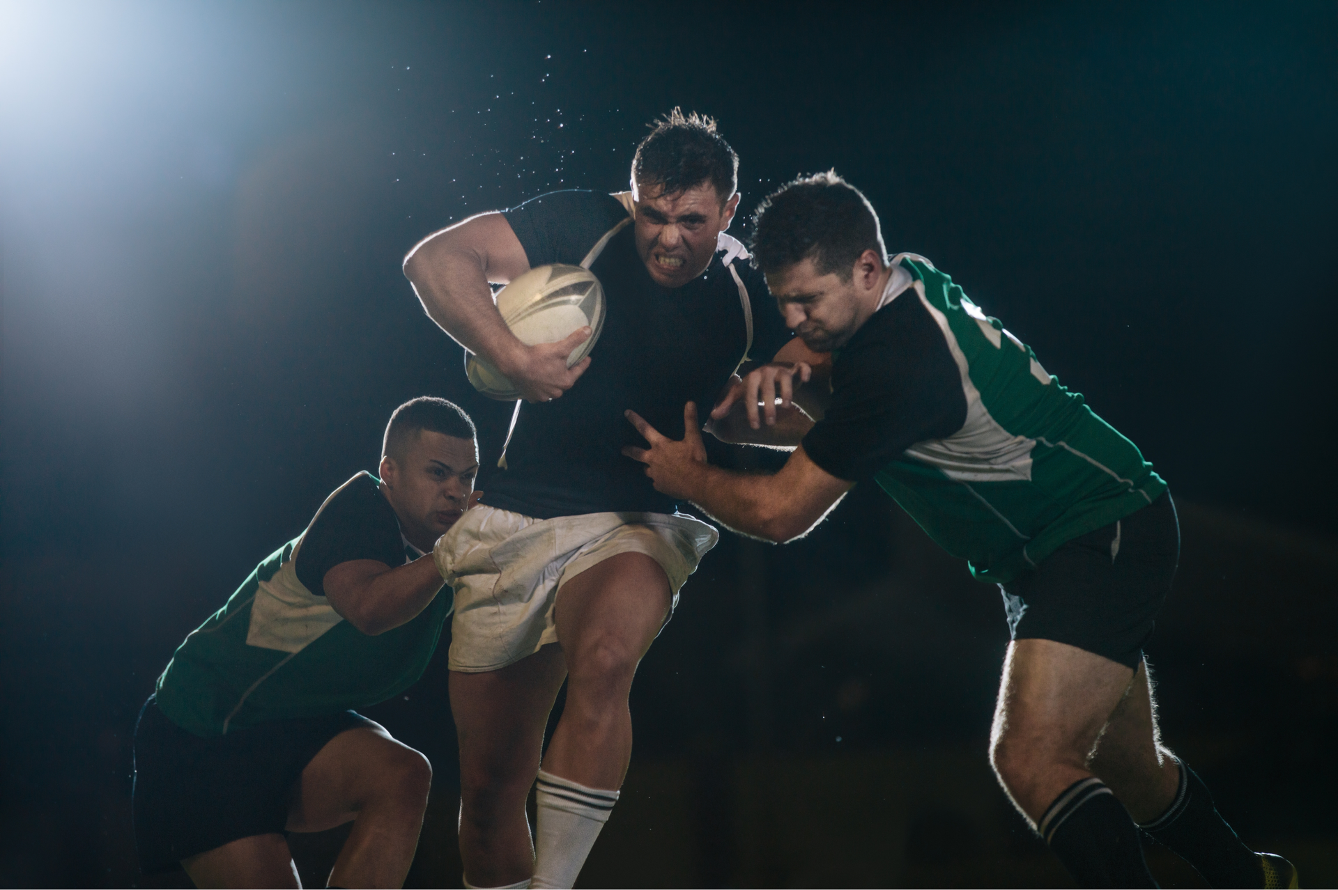 Rugby players in action; one running with ball, two tackling. Dark background, bright spotlight.