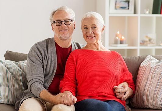 Elderly couple smiling, seated together on a couch indoors; man holds woman's hand.