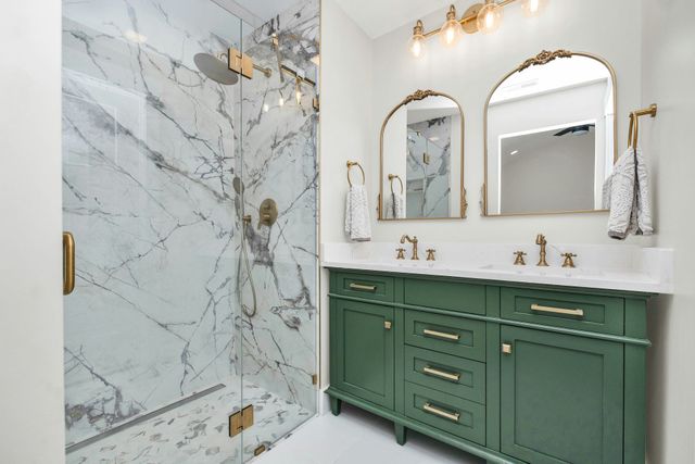 Green vanity bathroom with marble shower, gold fixtures, and arched mirrors.