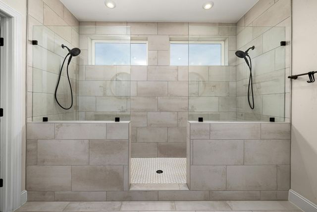 Bathroom with two glass-walled showers, light stone tile, black showerheads, and small windows above.