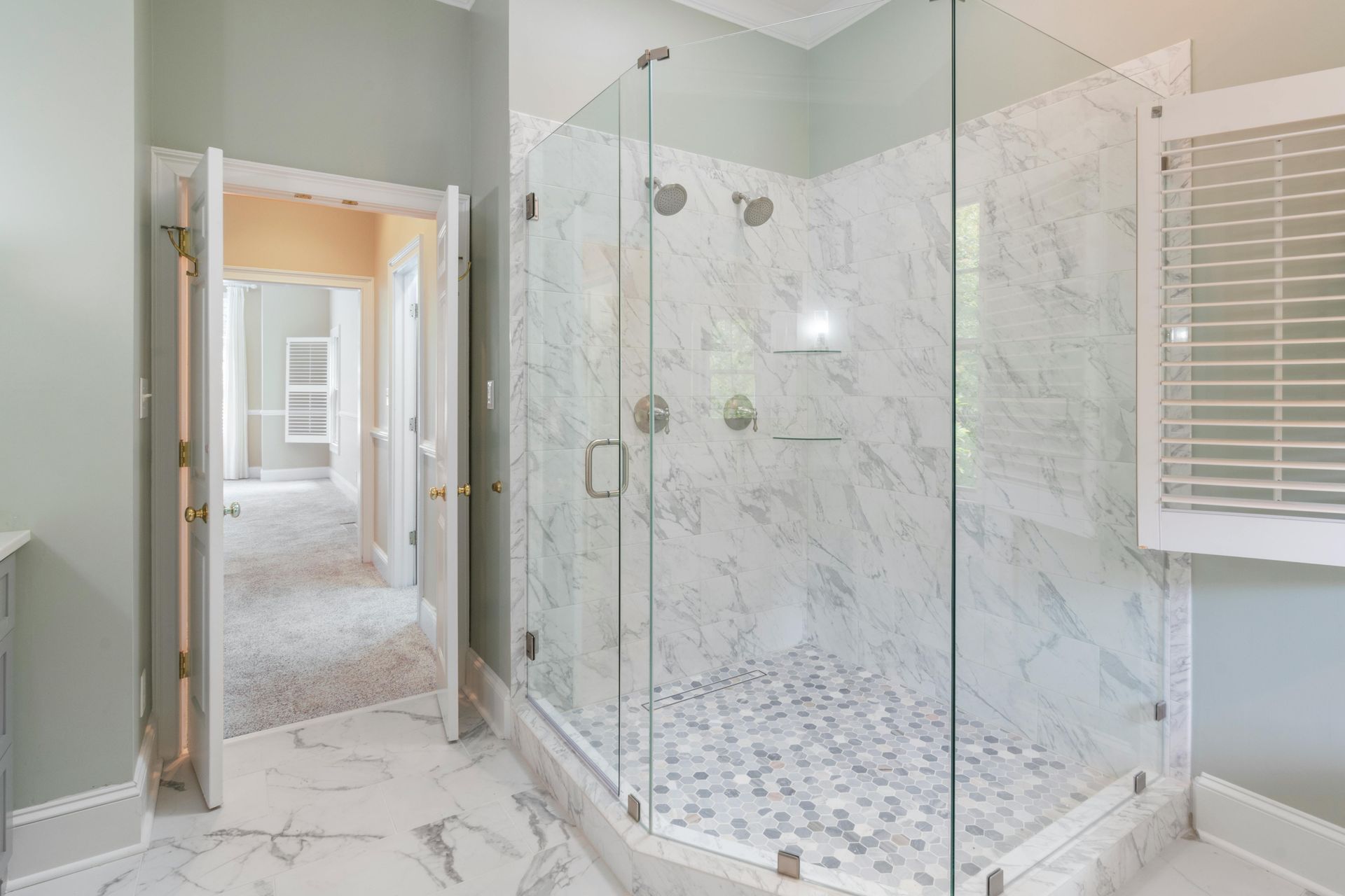 Spacious marble-tiled bathroom with a glass shower enclosure and a doorway leading to a hallway.