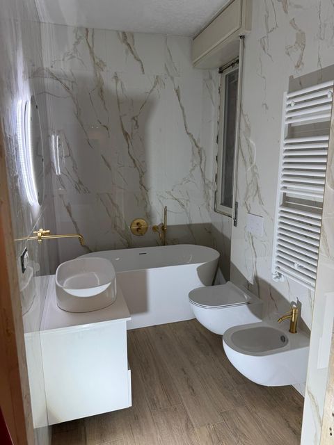 Un bagno moderno con vasca freestanding bianca, lavabo quadrato su mobiletto e un'ampia finestra con vista sugli alberi.